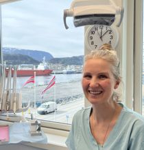 Smilende tannlege i blå skrubber, ved et vindu, med tannverktøy i bakgrunnen.