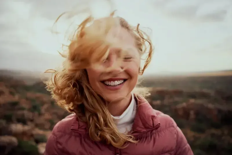 Kvinne med vindblåst hår smiler bredt utendørs, iført en rosa jakke mot et uskarpt landskap.