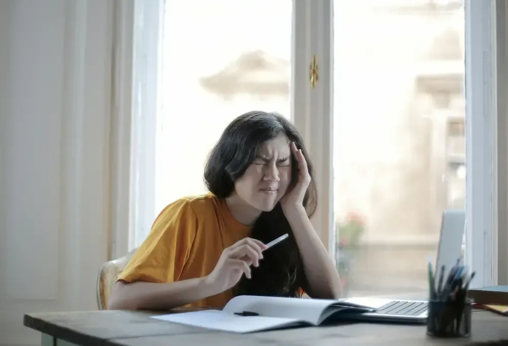 Kvinne med hodepine, holder penn, ved skrivebord med bærbar PC og notatbok.