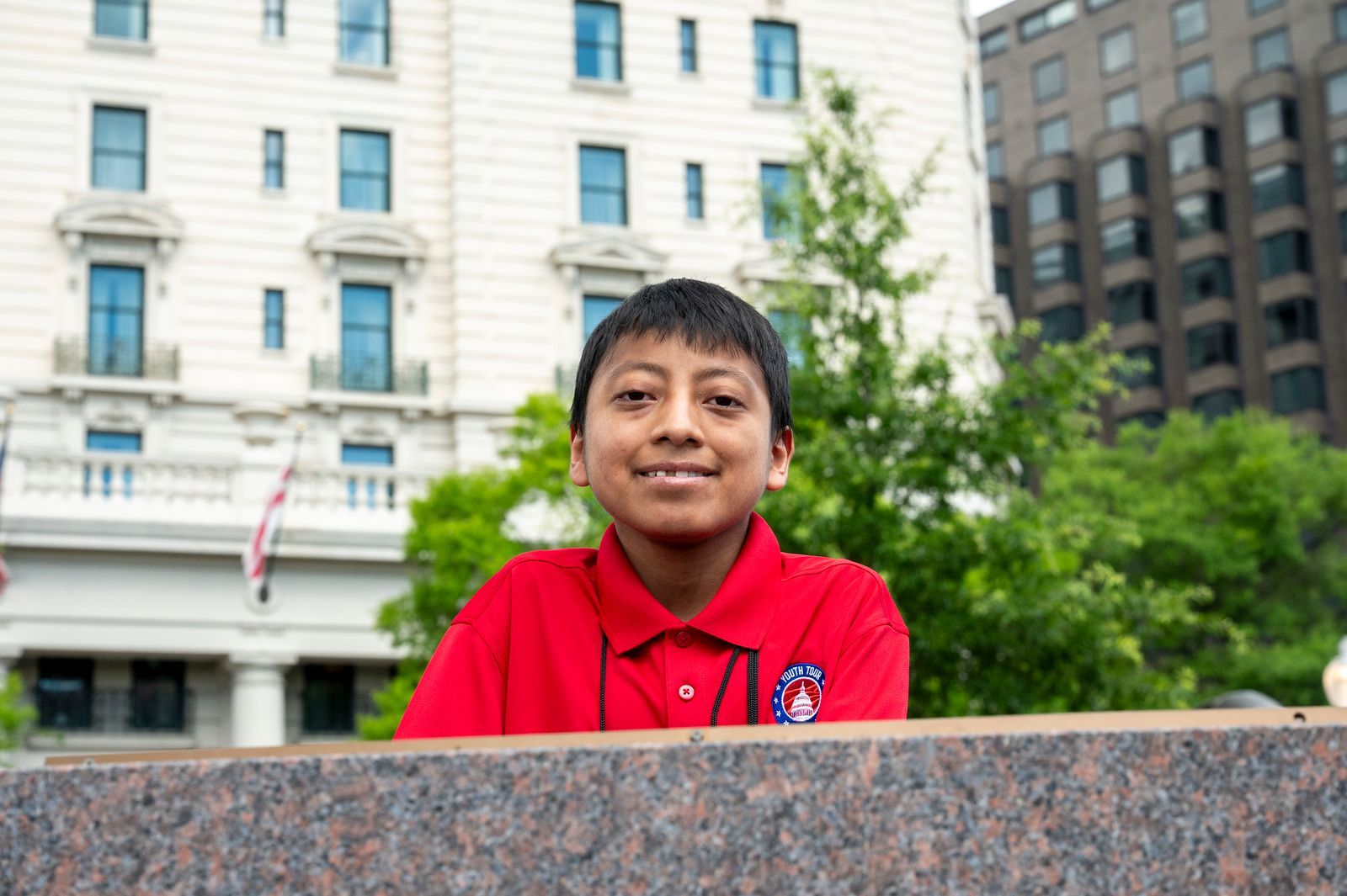 Person in a red shirt smiling outdoors in front of a building and trees