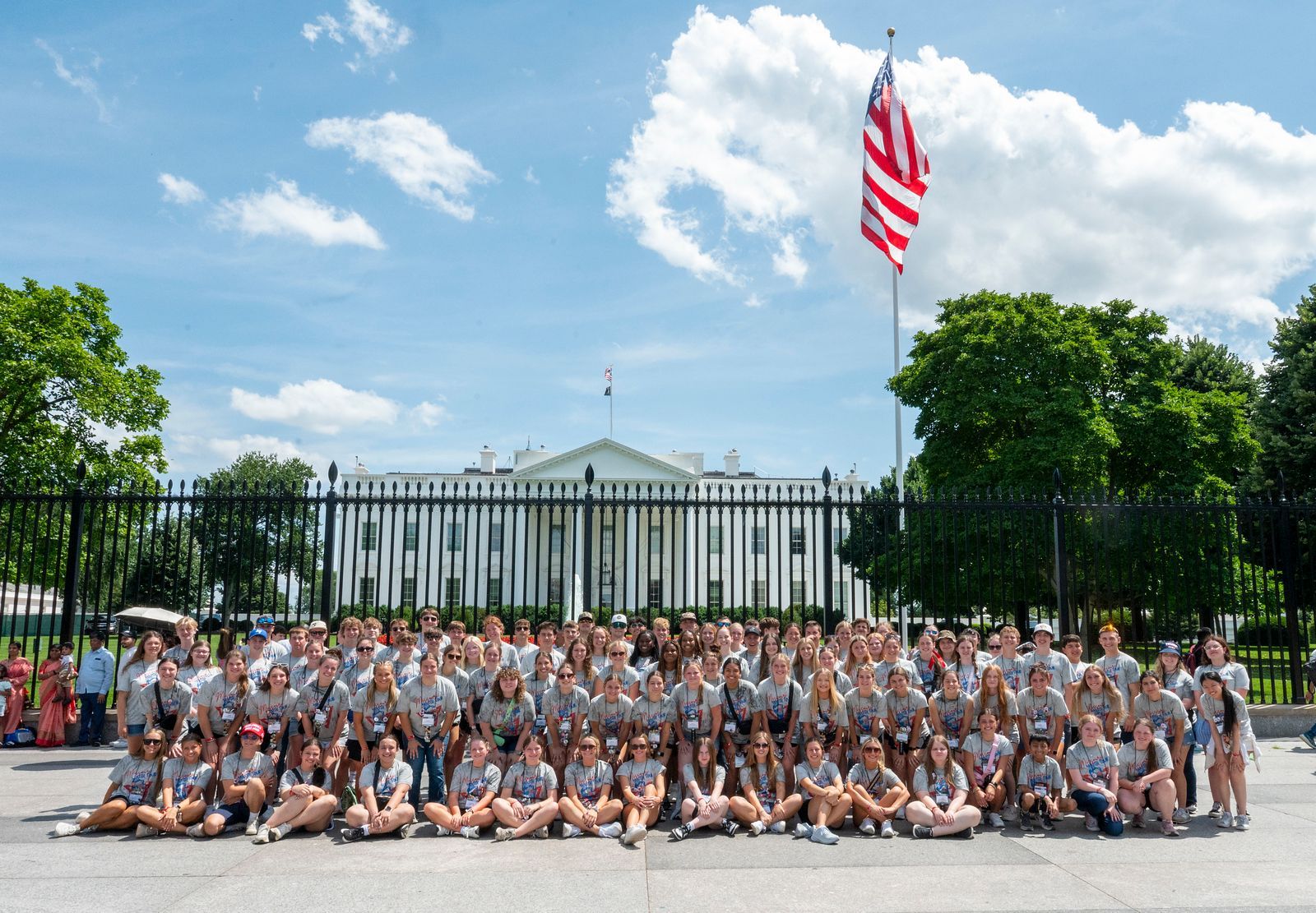 Large group posing in front of the White House with an American flag overhead under a sunny sky