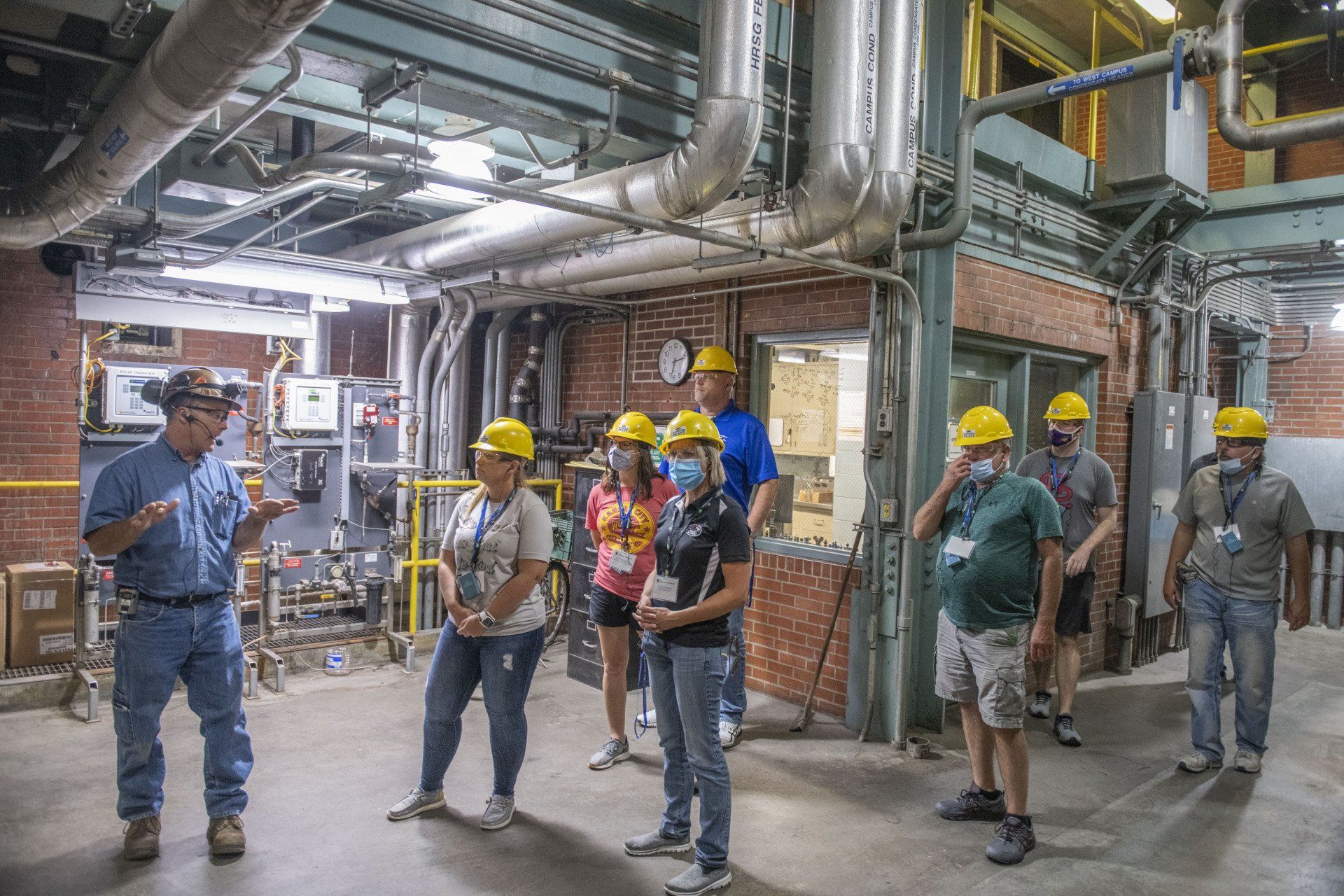 teachers in hard hats tour an industrial plant with pipes and machinery.