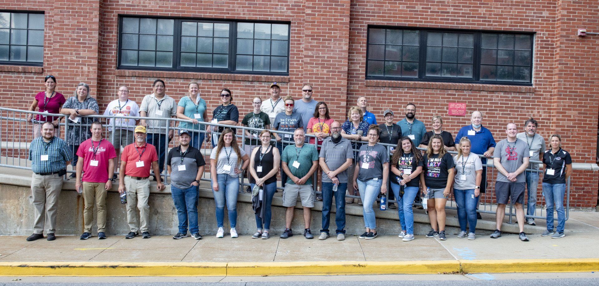 Large group posing for a photo outside a brick building, standing on a sidewalk and steps