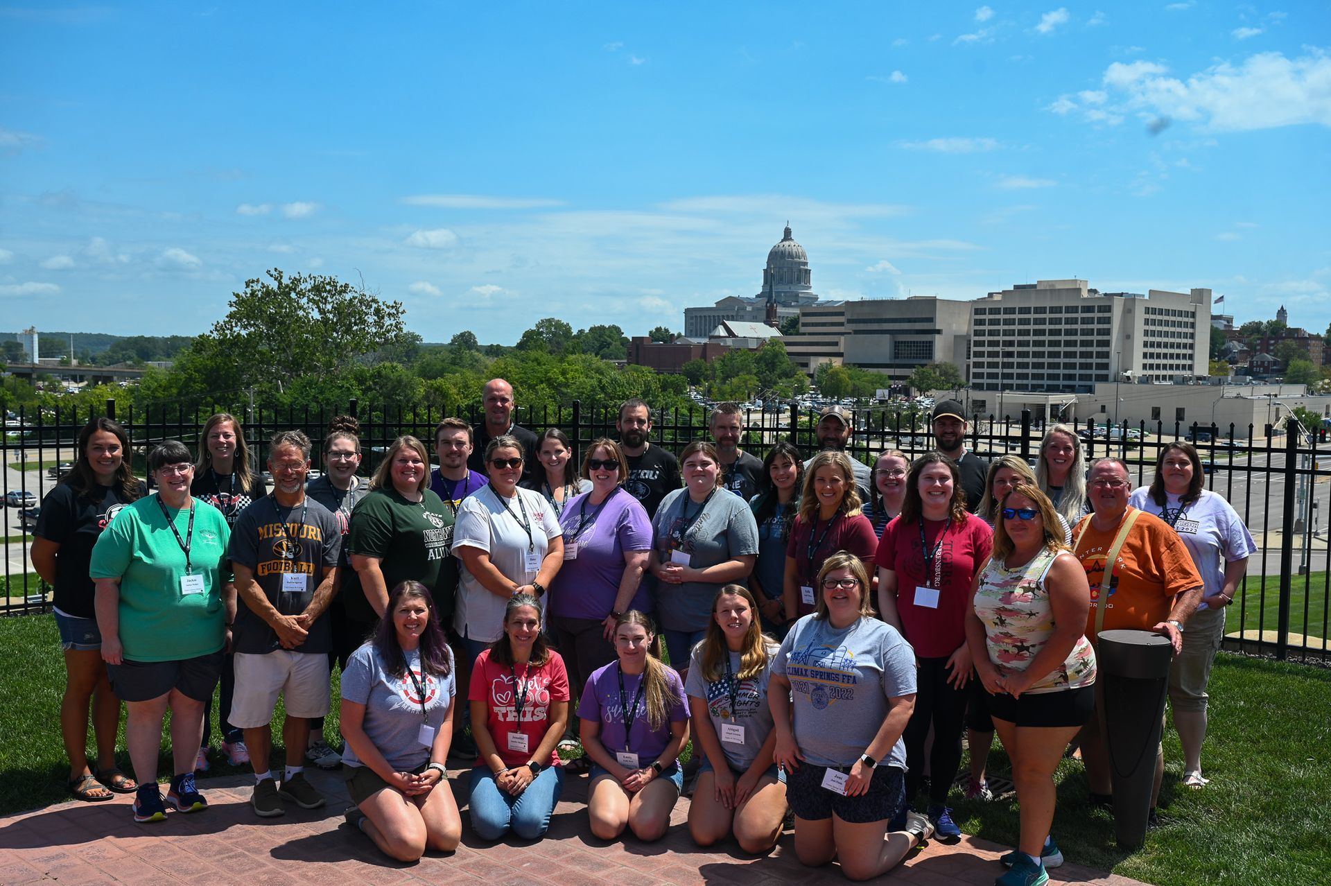 Large group posing outdoors in front of a city skyline and brick plaza on a sunny day