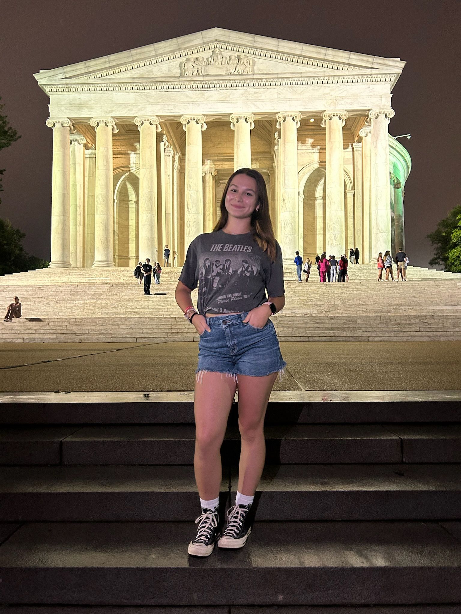 Woman standing on steps at night in front of a brightly lit neoclassical monument.