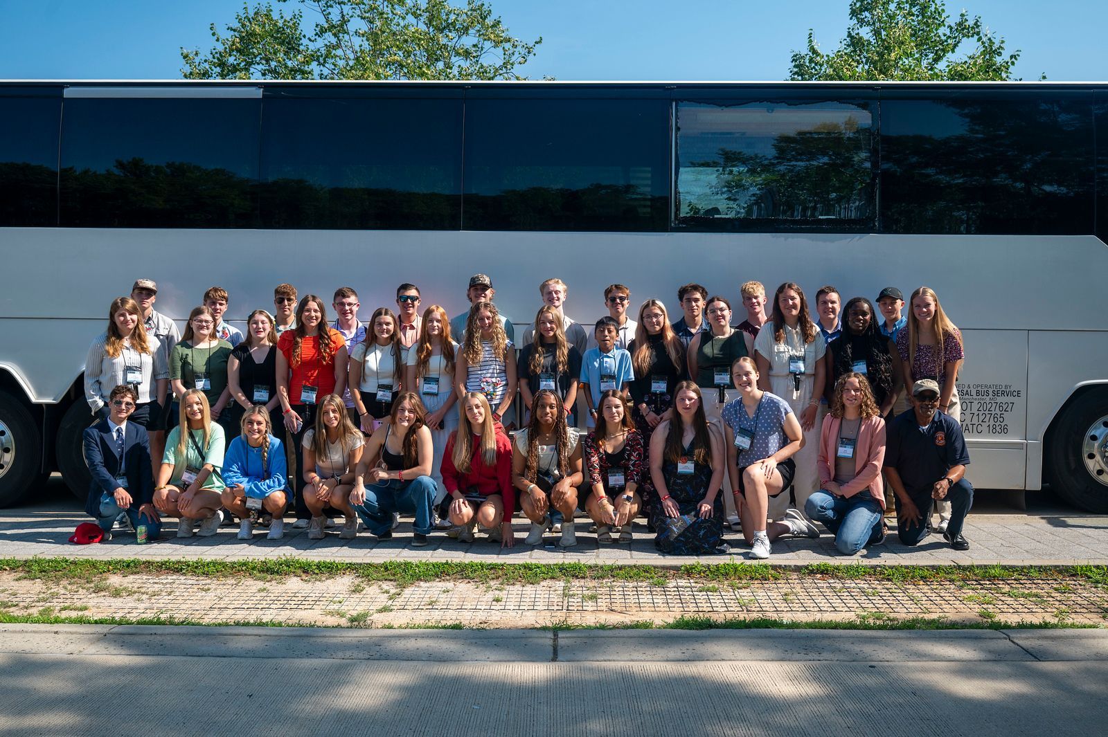 Large group posing in front of a white bus on a sunny street, with trees and grass behind them.