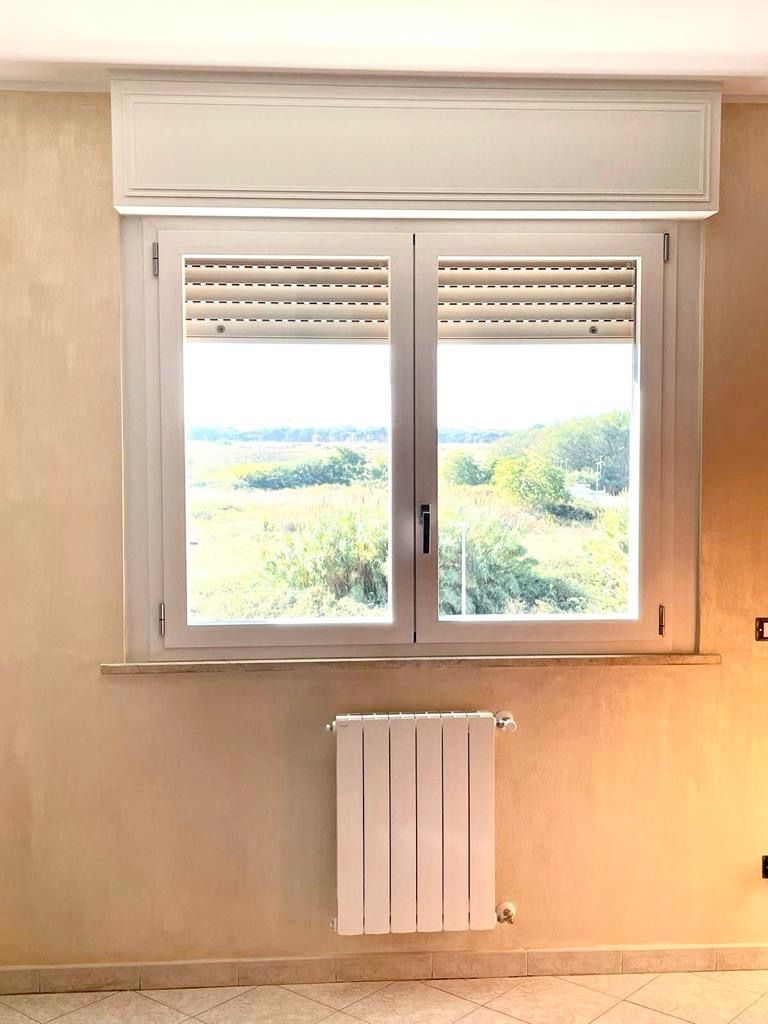 Finestra con cornice bianca, tapparella e termosifone sottostante, affacciata su un paesaggio verdeggiante.