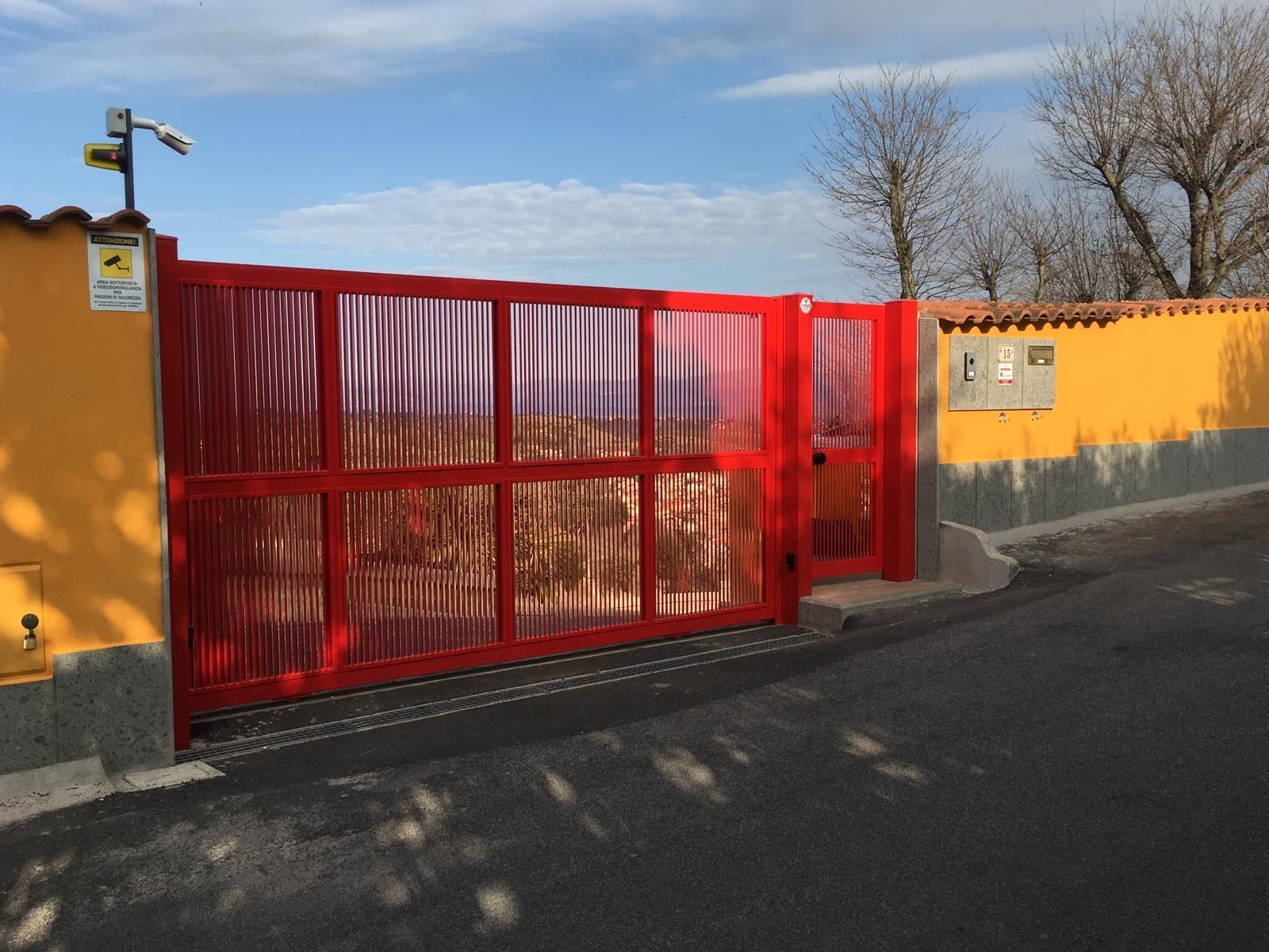 Un cancello di metallo rosso incastonato in un muro giallo, accanto a una strada asfaltata, sotto un cielo azzurro.