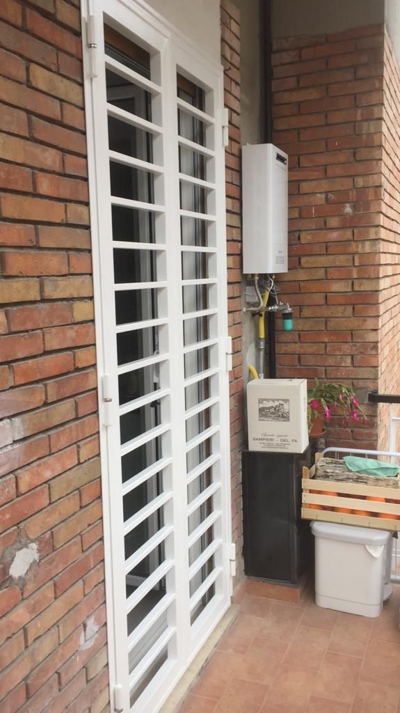 Balcone in mattoni con porta blindata bianca, scatola di derivazione e oggetti impilati accanto a un muro.