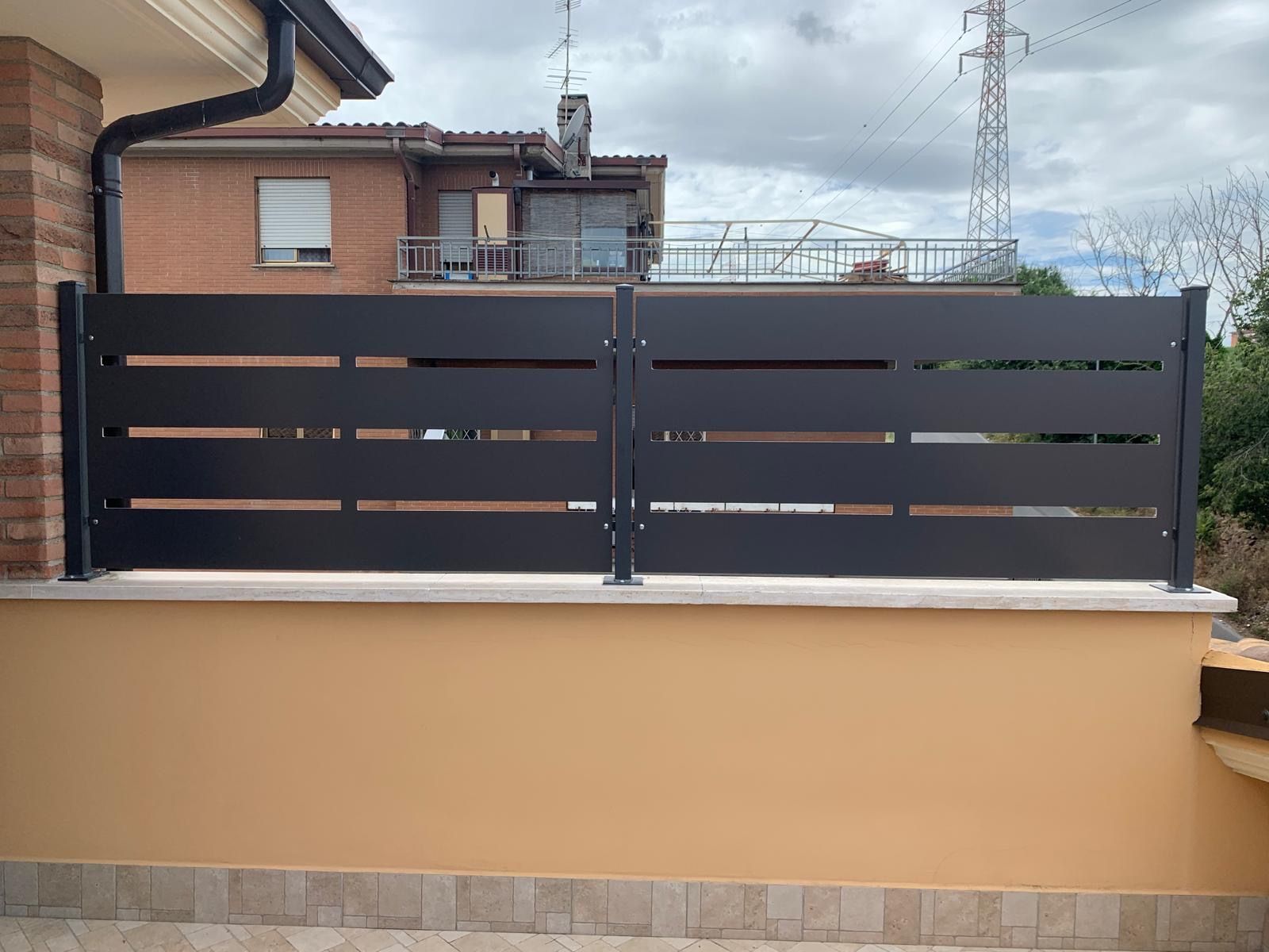 Cancello in metallo a doghe grigio scuro sopra un muro beige di fronte a una casa di mattoni sotto un cielo nuvoloso.