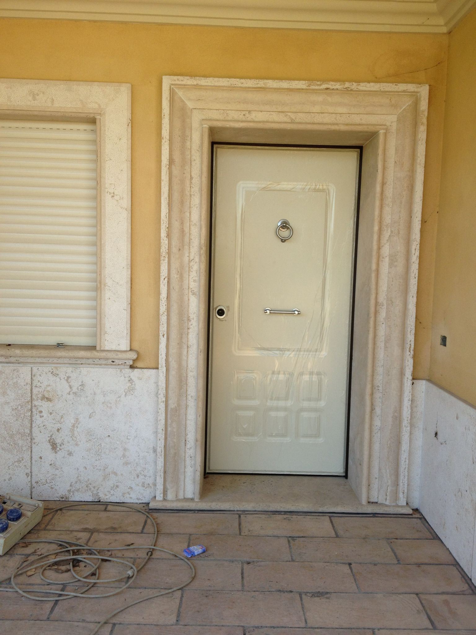 Porta d'ingresso bianca chiusa con finiture beige e finestra con persiane su un portico in pietra consumata