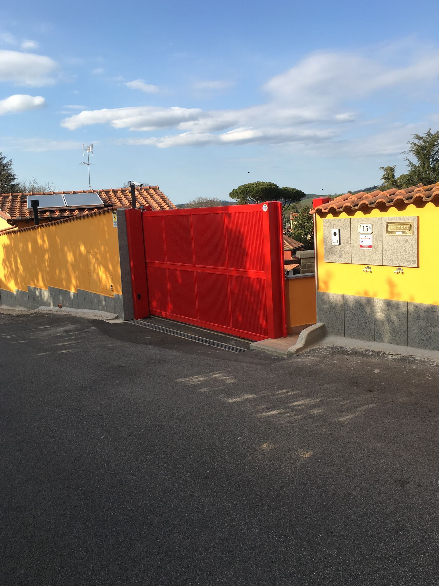 Muri gialli e un cancello rosso accanto a una strada asfaltata sotto un cielo azzurro con nuvole