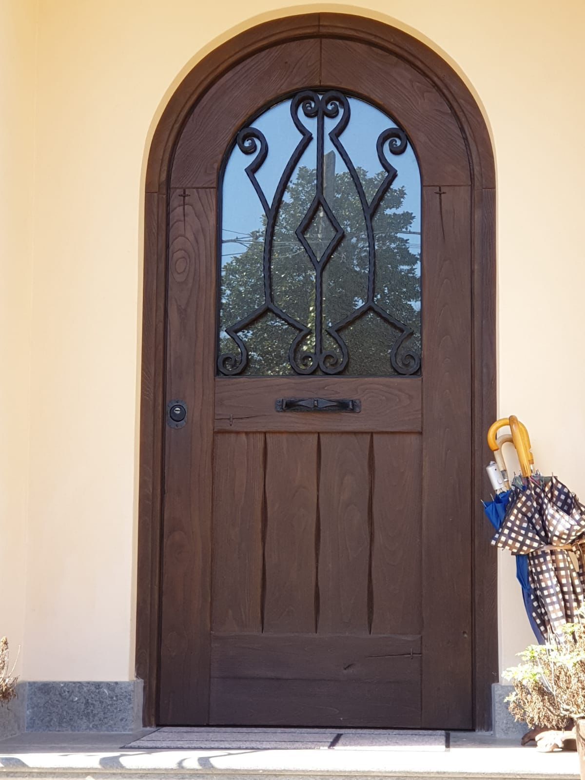 Porta d'ingresso ad arco marrone con pannello in vetro e ferro battuto accanto a una parete color crema e portaombrelli.