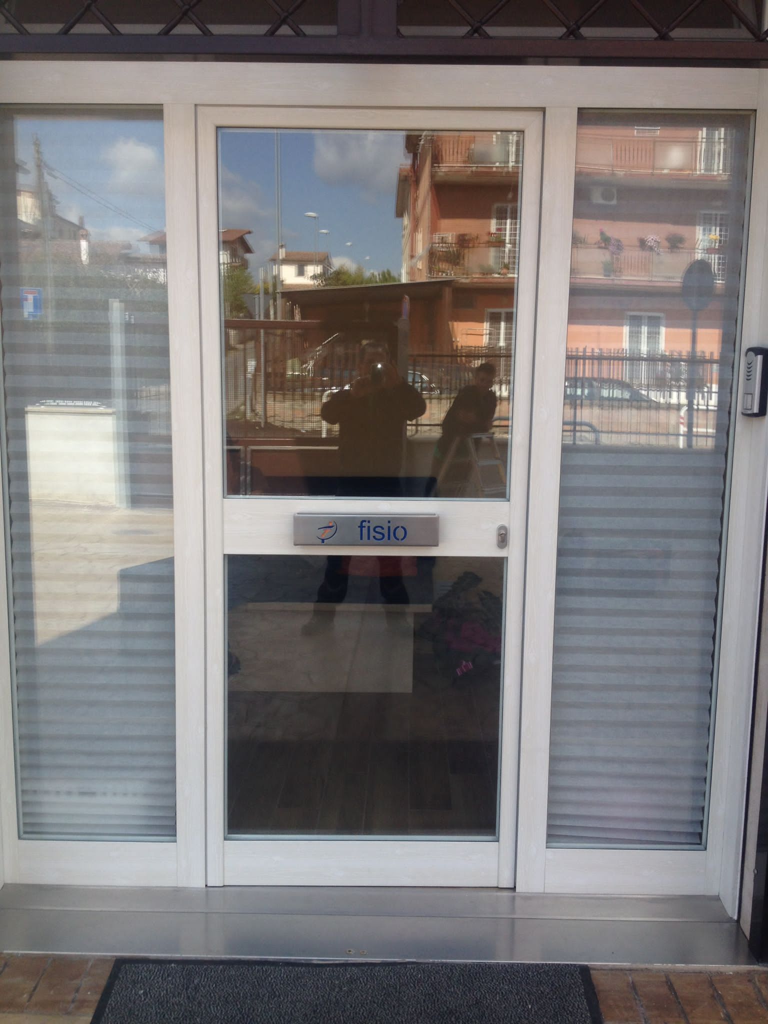 Ingresso del negozio con vetrina a vetri, doppie porte e persiane metalliche su entrambi i lati, che riflettono gli edifici circostanti.