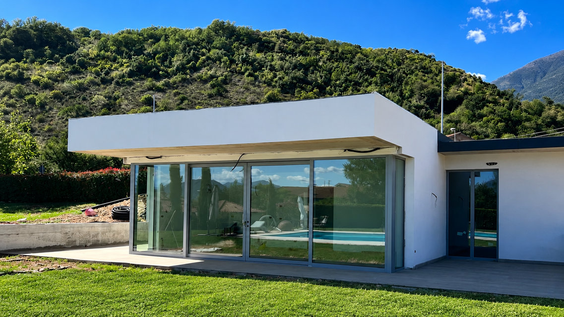 Casa moderna bianca con pareti in vetro e tetto piano, immersa nel verde con colline e montagne sullo sfondo.