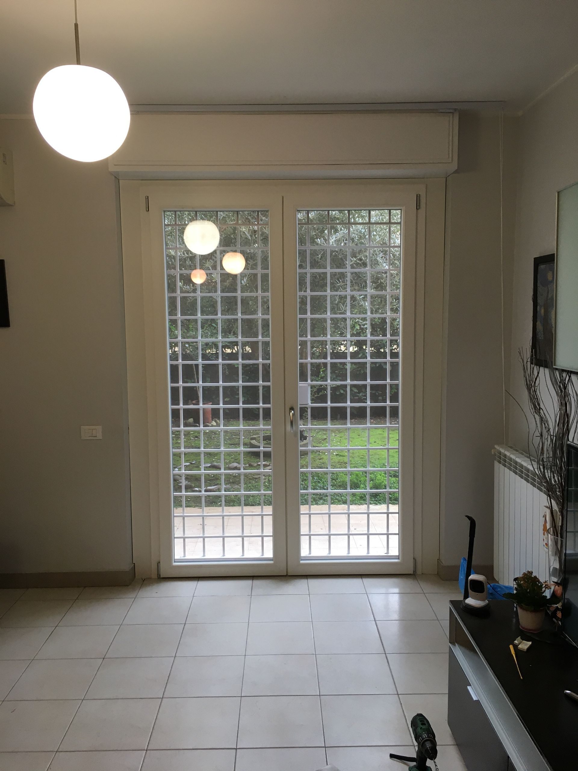 Soggiorno vuoto con pavimento piastrellato, doppie porte a vetri e una lampada rotonda sospesa vicino al soffitto.