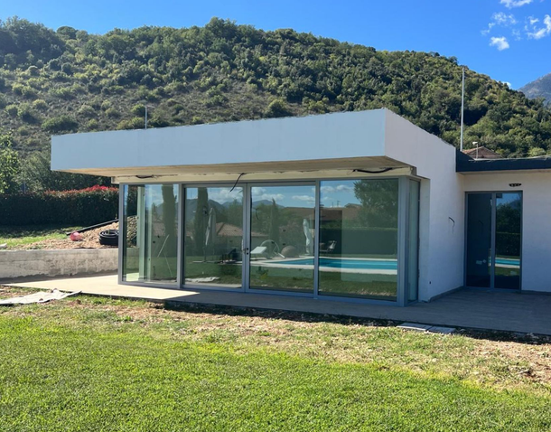 Moderno edificio con facciata in vetro e tettoia bianca, affacciato su un prato verde e colline verdi sotto un cielo azzurro.