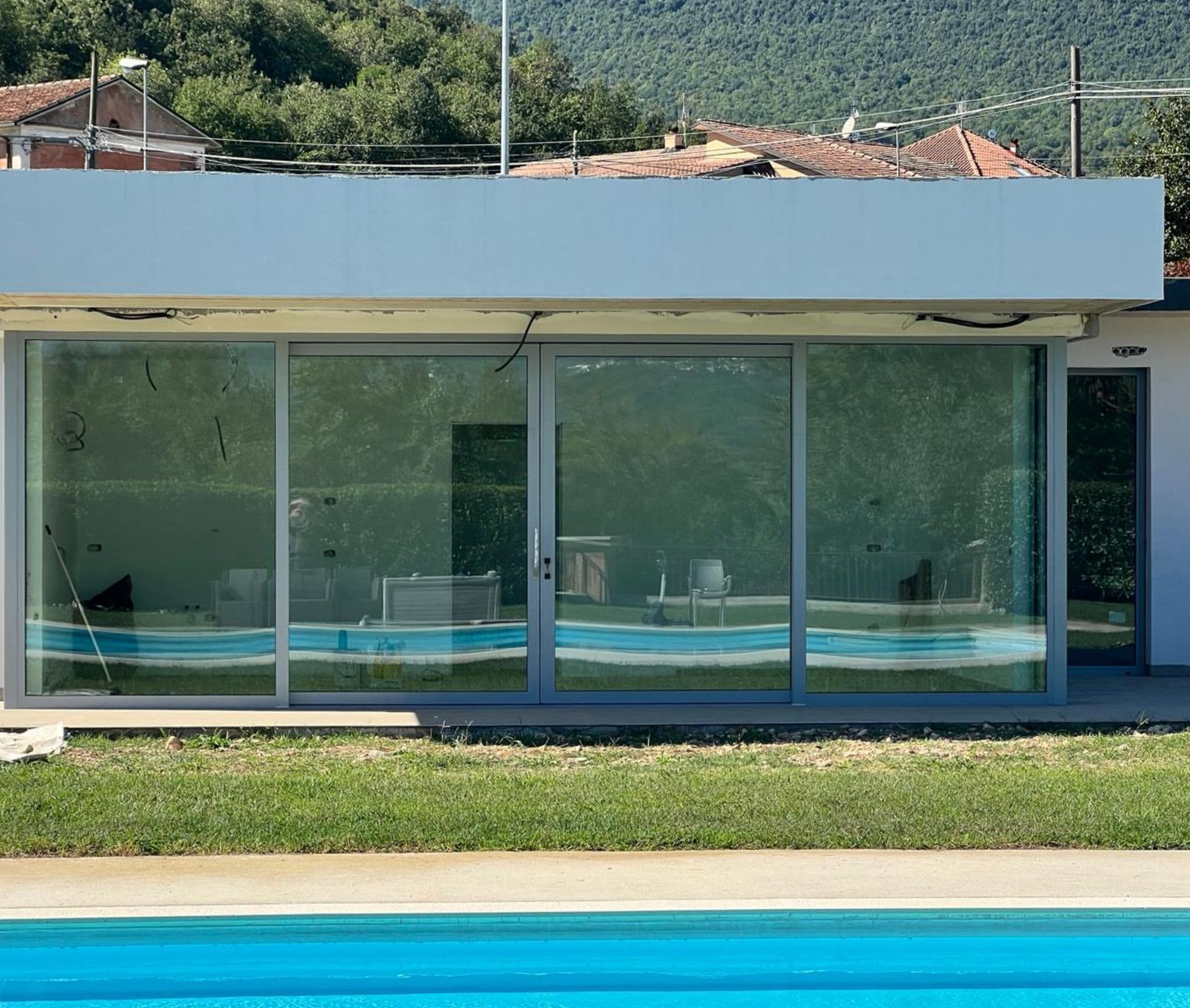 Casa moderna con facciata in vetro, piscina antistante e colline sullo sfondo.
