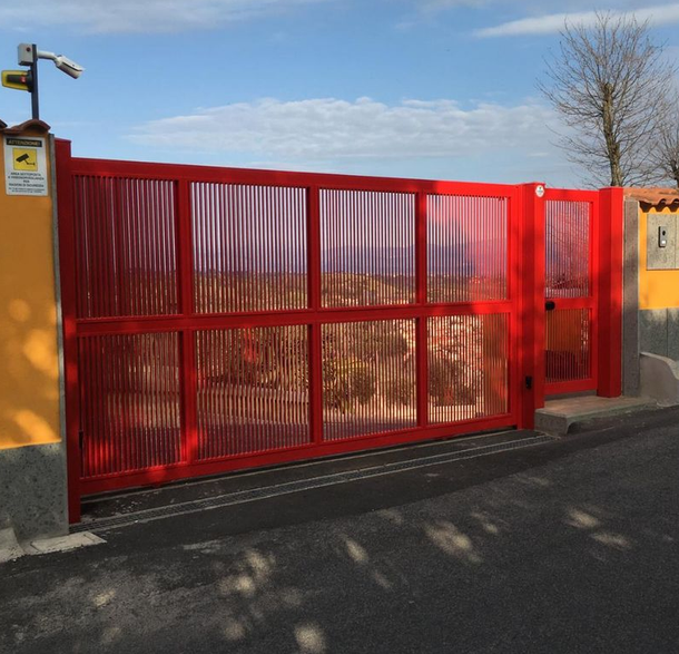 Un cancello scorrevole in metallo rosso accanto a muri gialli sul ciglio della strada, con una telecamera di sicurezza sopra.