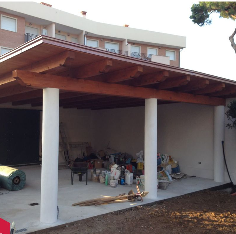 Posto auto coperto con tetto in legno e pilastri bianchi accanto a una casa, ingombrato da oggetti riposti al di sotto.