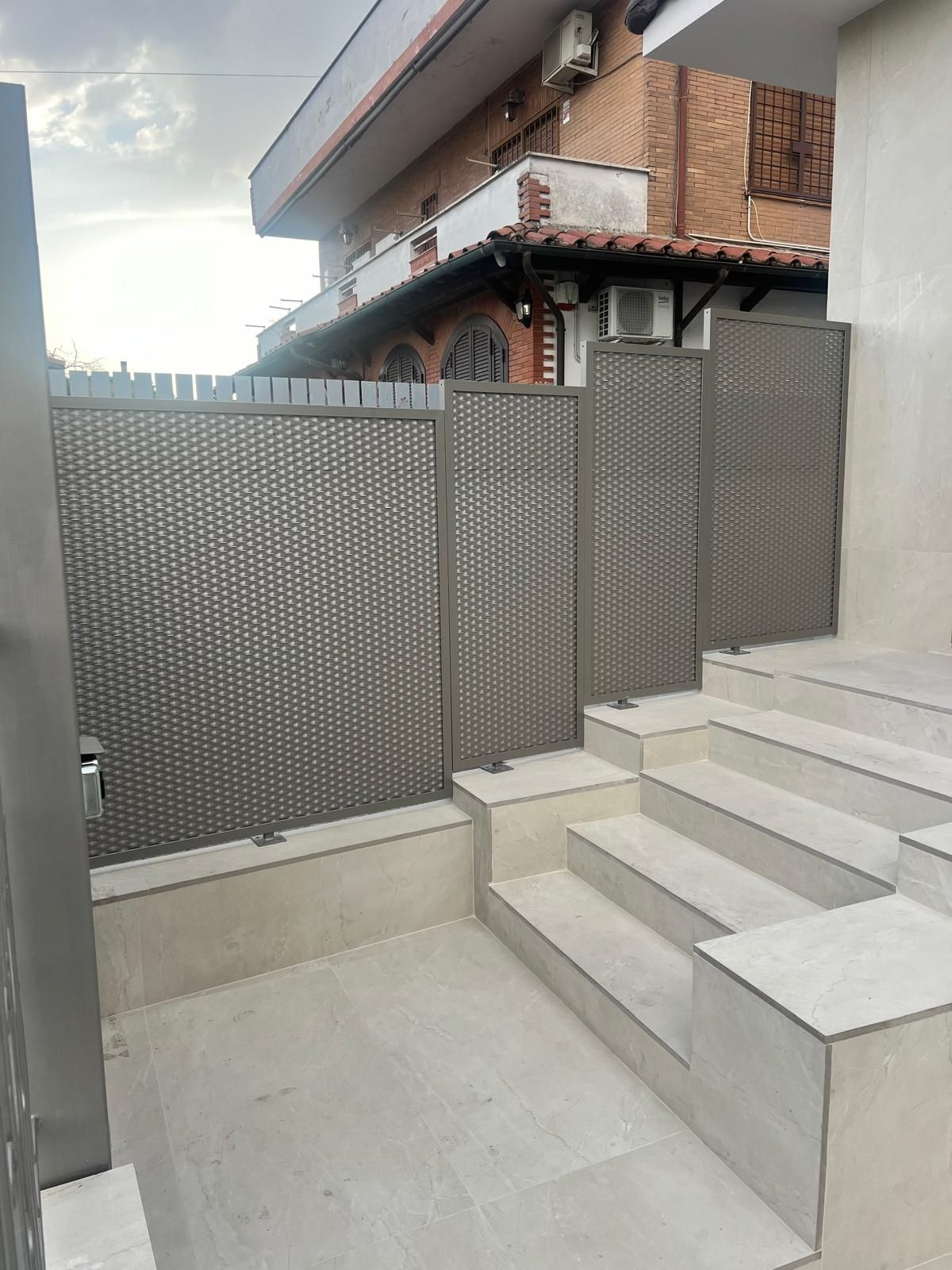 Scalini in cemento accanto a una recinzione metallica grigia e al muro del patio in un cortile esterno.