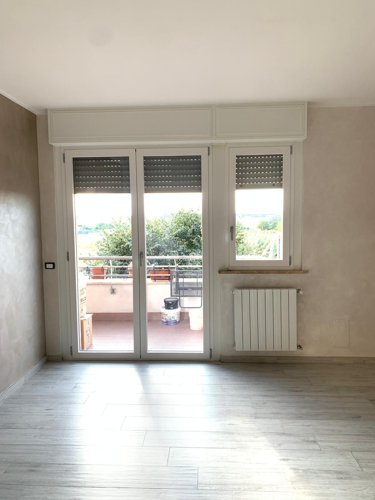 Soggiorno vuoto con porte scorrevoli in vetro che si aprono su un balcone e termosifone sotto la finestra.