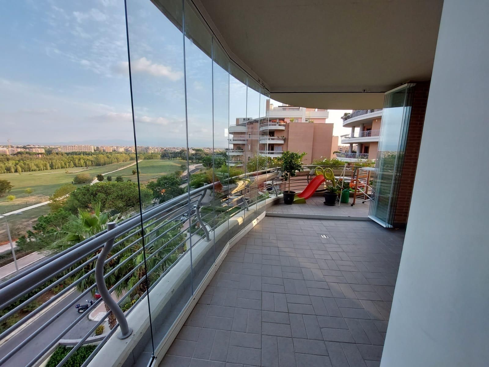 Balcone affacciato su una strada e sul verde, con ringhiera in vetro e palazzi residenziali in lontananza.