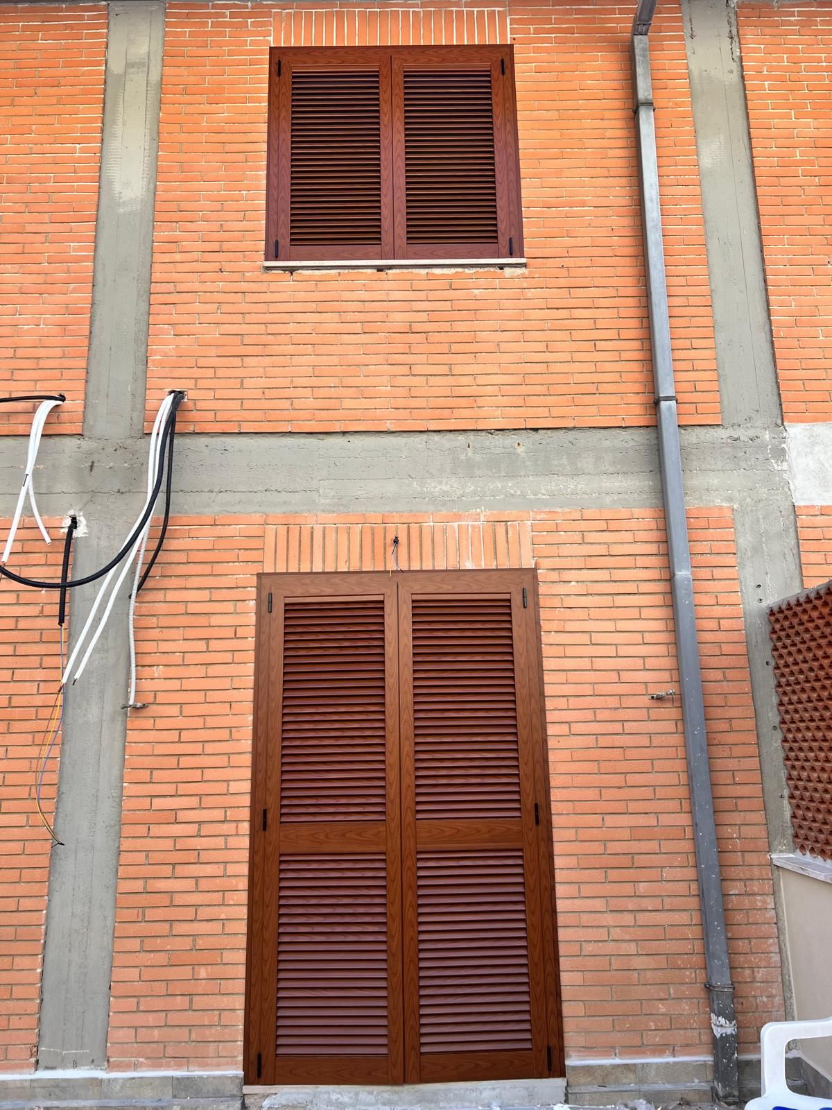 Facciata dell'edificio in mattoni con fasce di cemento grigio, due finestre a persiana marroni e una porta.