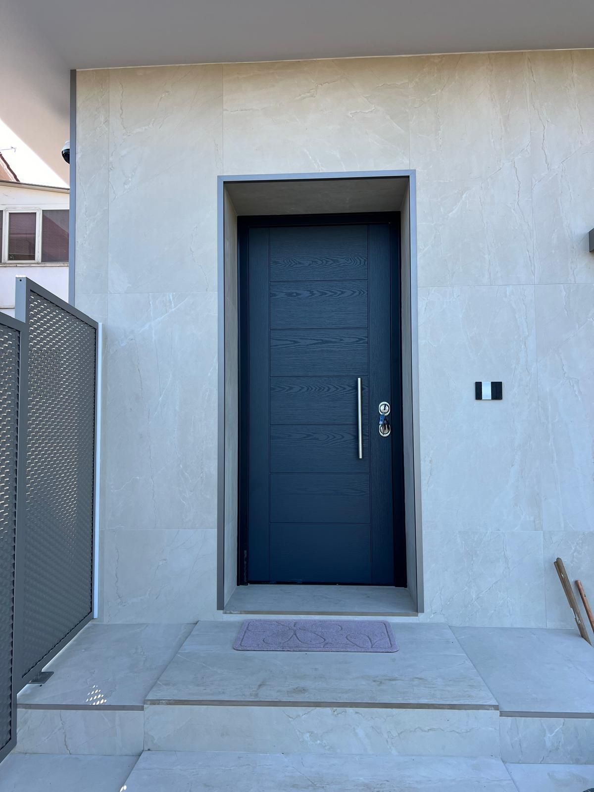 Ingresso principale con porta blu scuro incastonata in un muro di stucco, scalini in cemento e cancello metallico sulla sinistra.