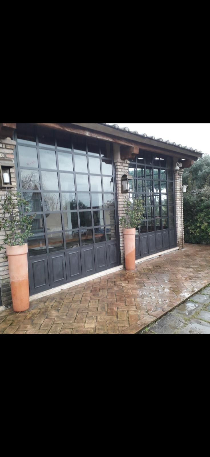 Patio con facciata in vetro, griglie metalliche, colonne con vasi e un vialetto in mattoni bagnati di fronte a una casa.