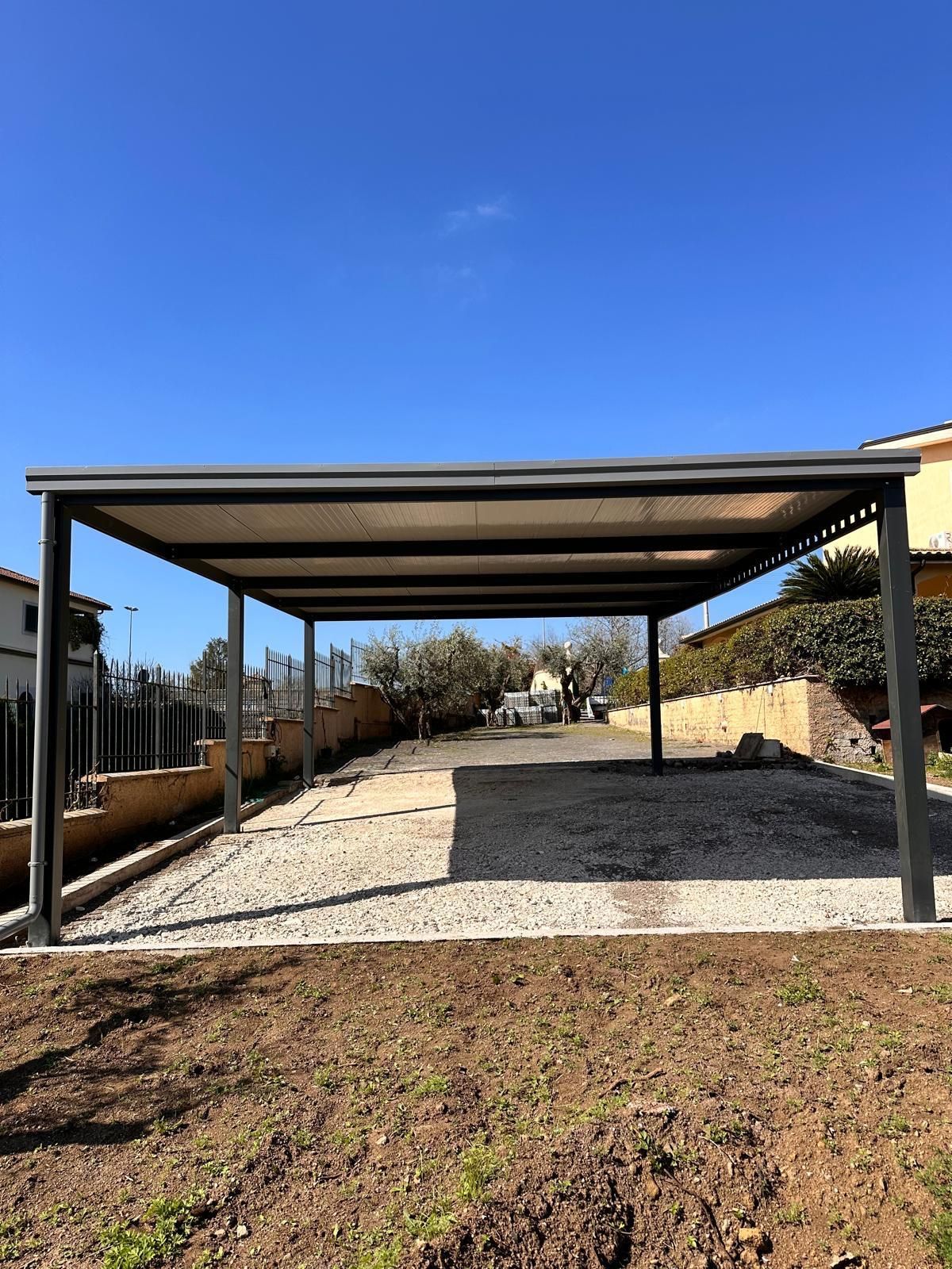 Posto auto coperto in metallo all'aperto su un terreno ghiaioso sotto un cielo azzurro e limpido, con un muro di pietra e alberi nelle vicinanze.