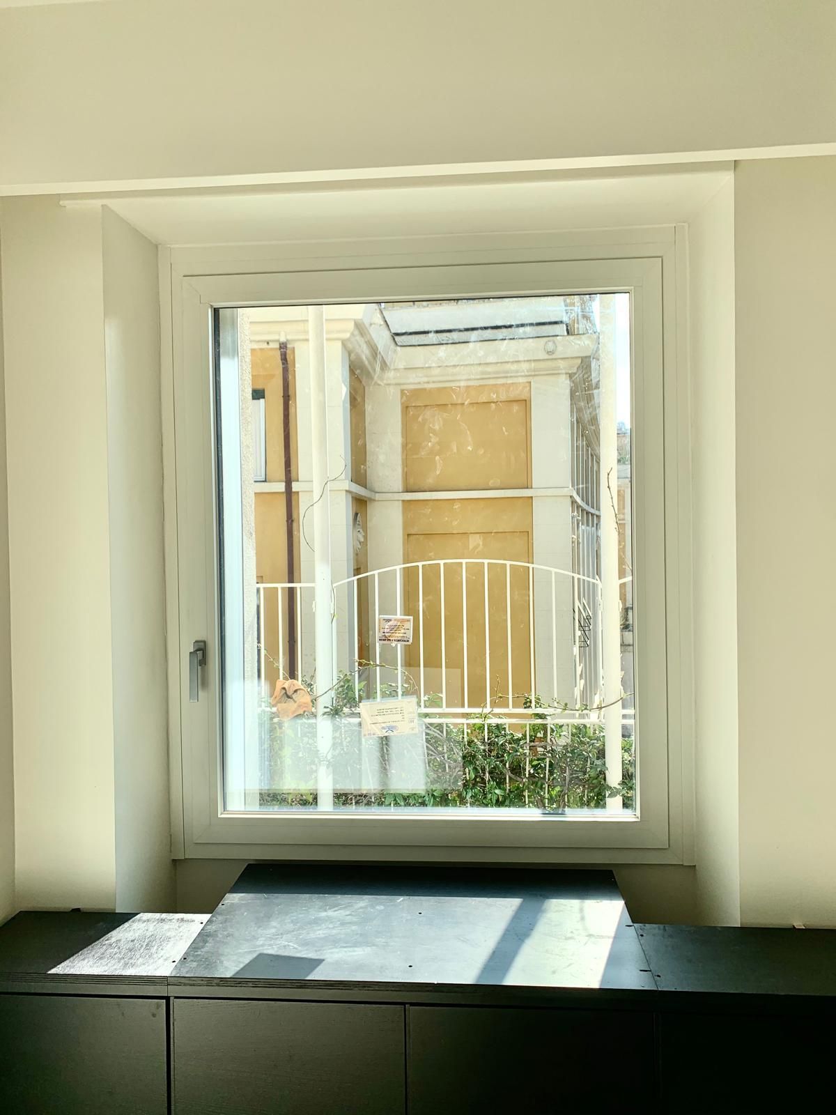 Finestra soleggiata che si affaccia su un balcone e sull'edificio adiacente, con una panchina scura in primo piano.
