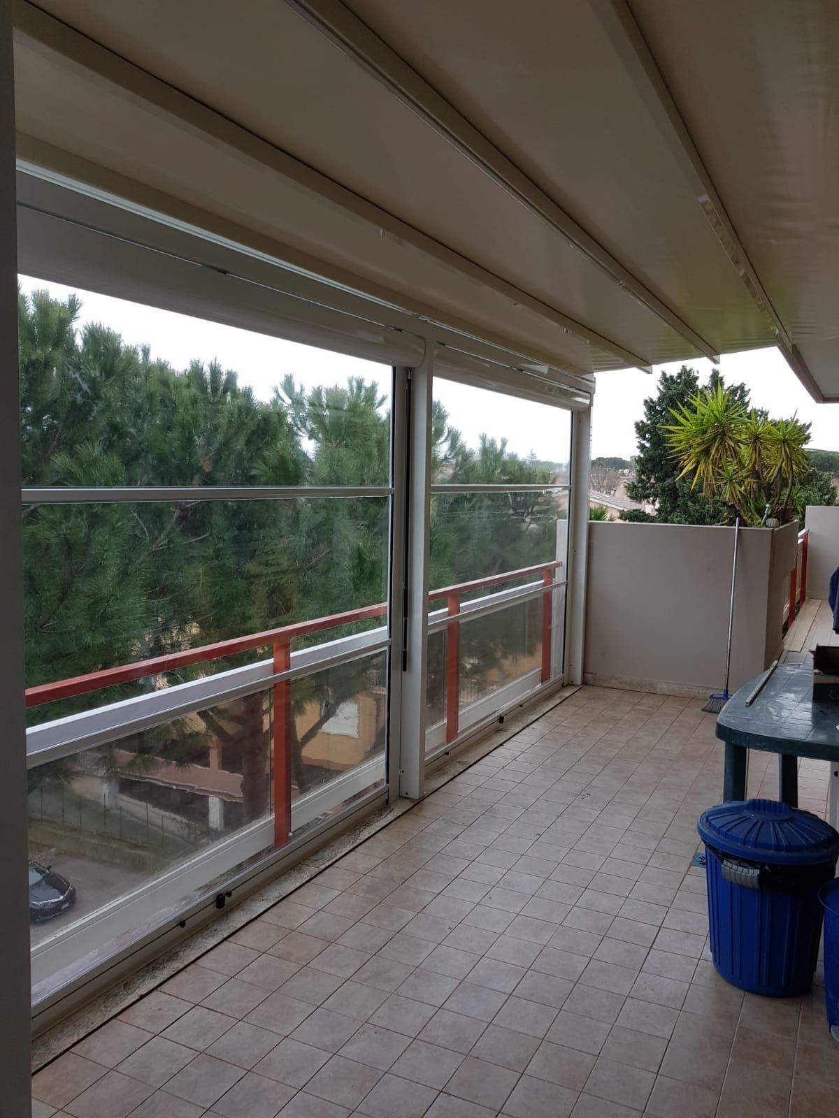 Balcone coperto con ringhiera in vetro, pavimento piastrellato, tavolo, cestino blu e pianta in vaso con vista sugli alberi.