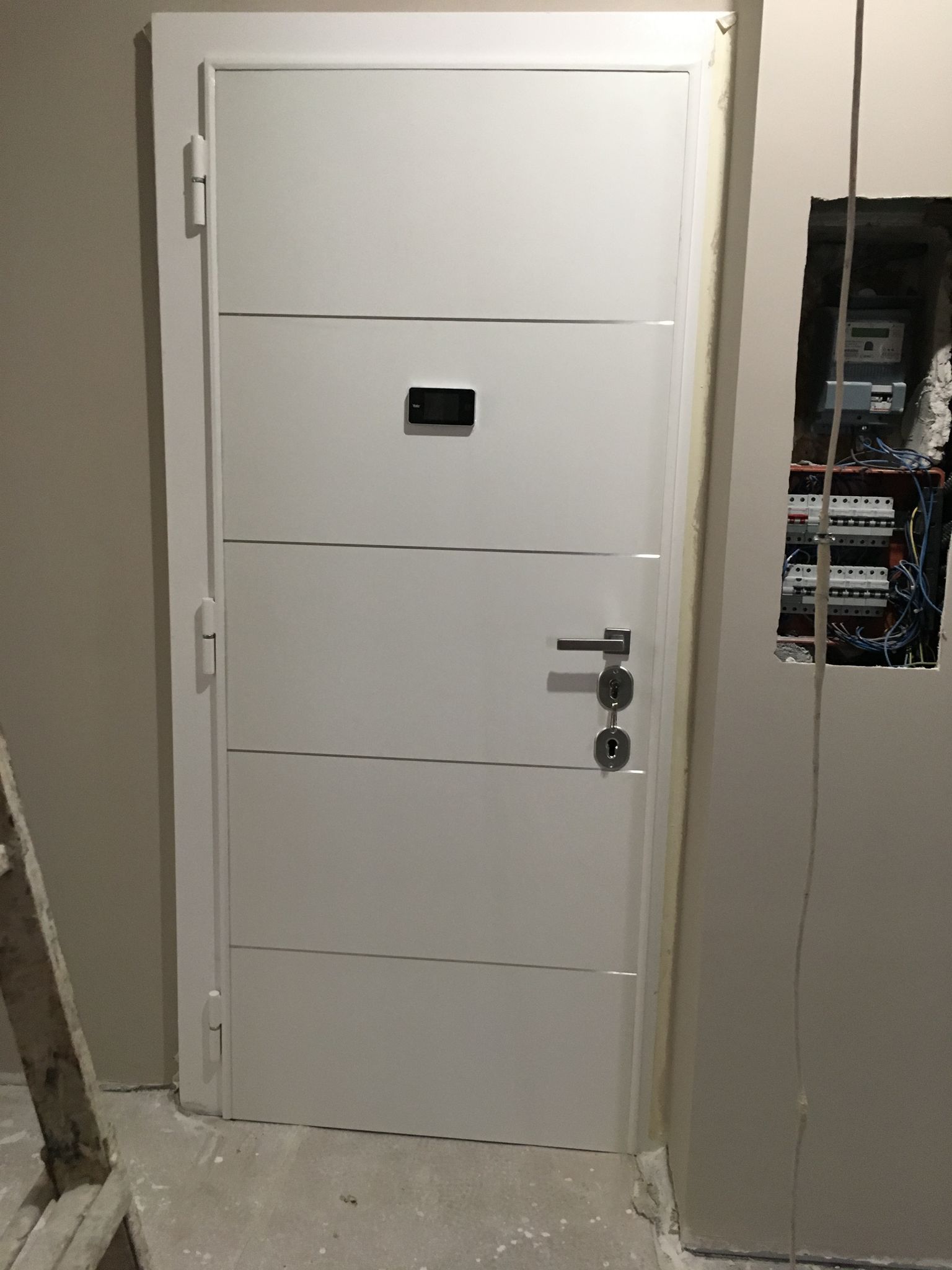 Porta metallica bianca con una piccola finestra in una parete interna beige, accanto ai pannelli elettrici.