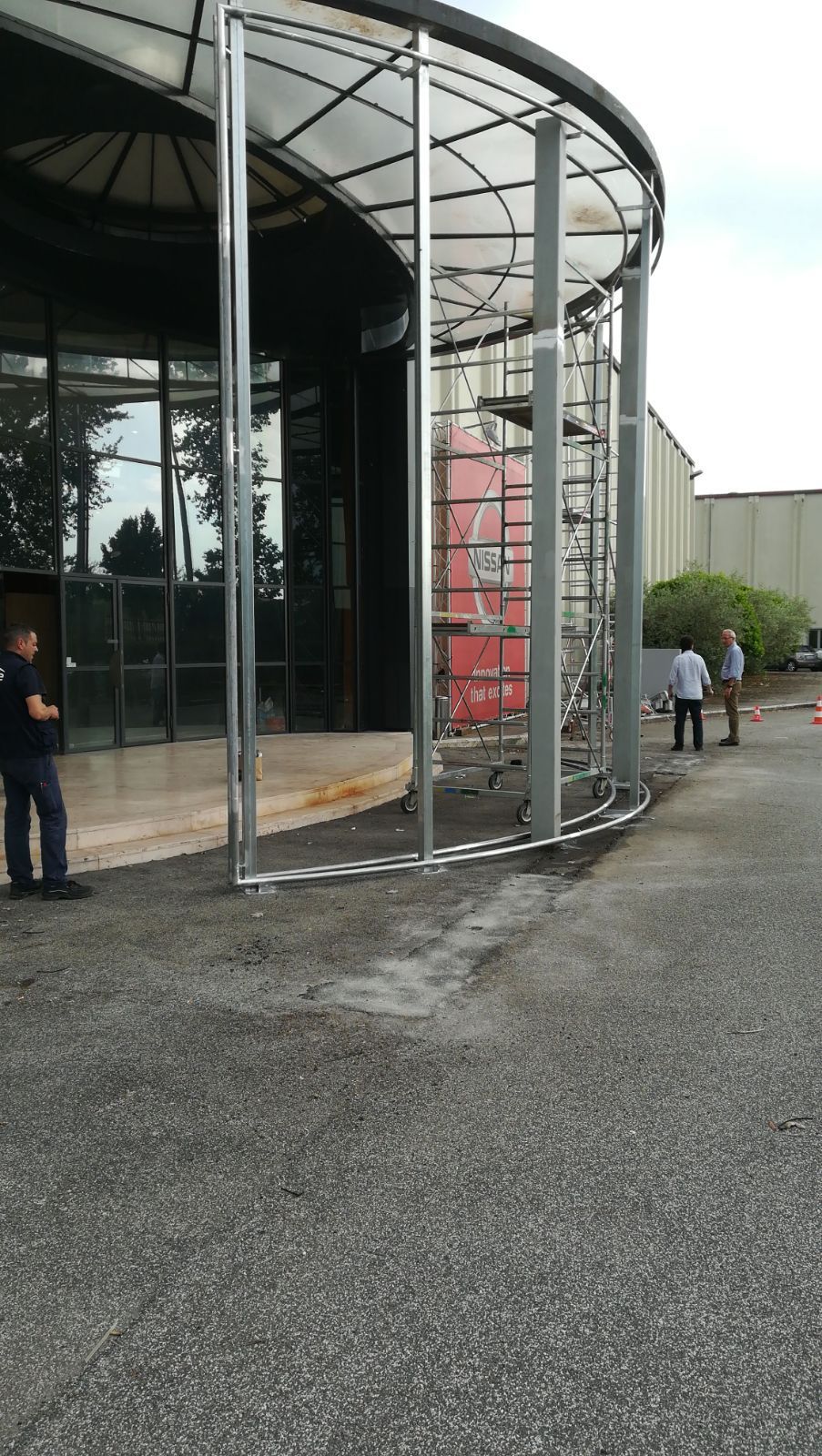 Ingresso con porta girevole in vetro accanto a un sentiero di ghiaia, con alcune persone in piedi nelle vicinanze.