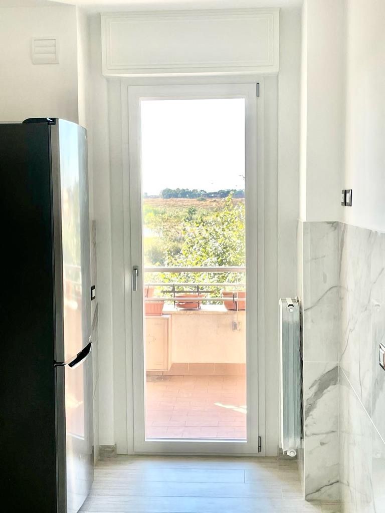 Cucina luminosa con frigorifero in acciaio inox, mobili bianchi, parete in marmo e porta a vetri che dà su un balcone con piante.