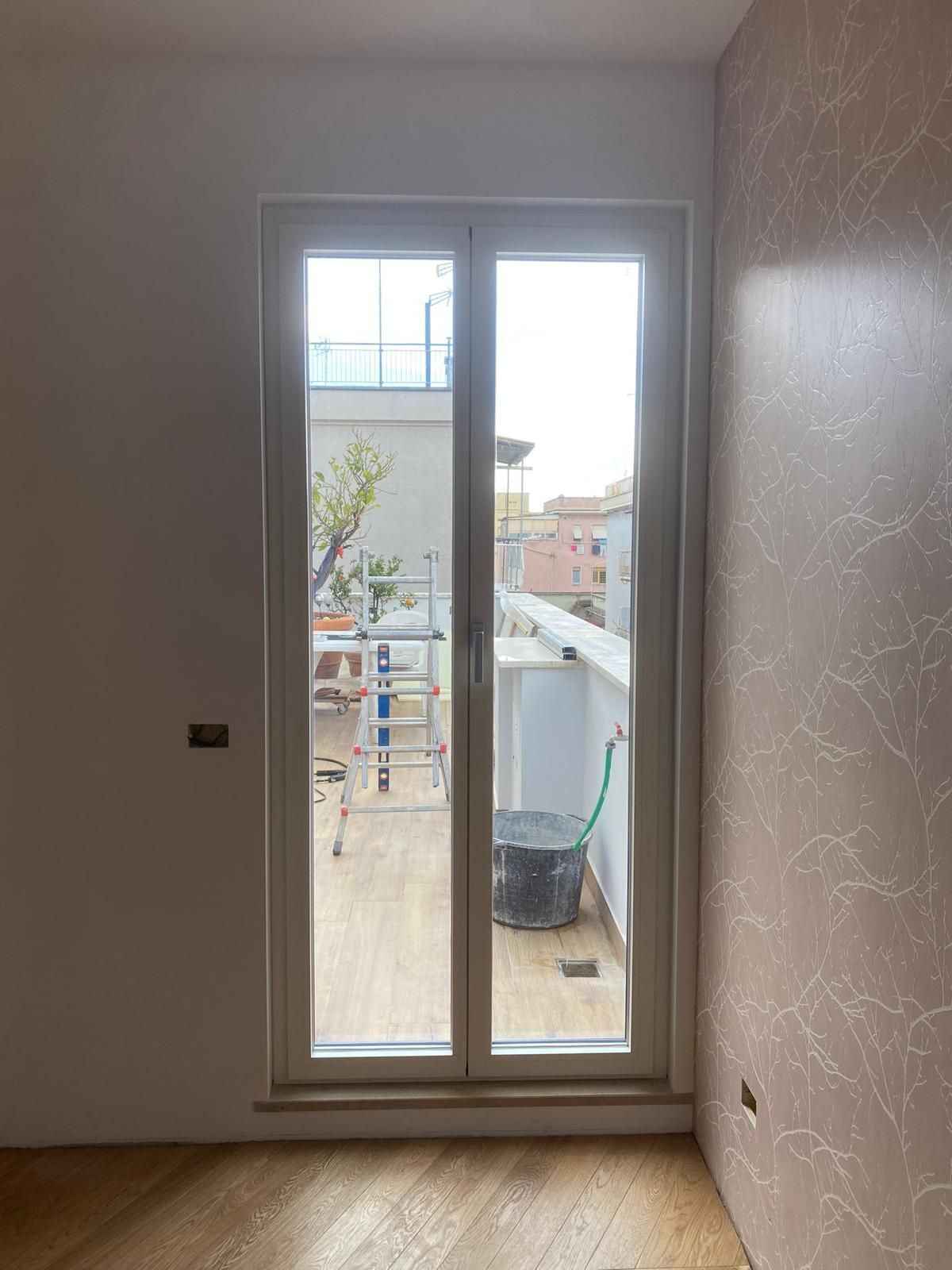 Vista interna attraverso una porta a vetri su un balcone con scala, scopa e, sullo sfondo, edifici della città.