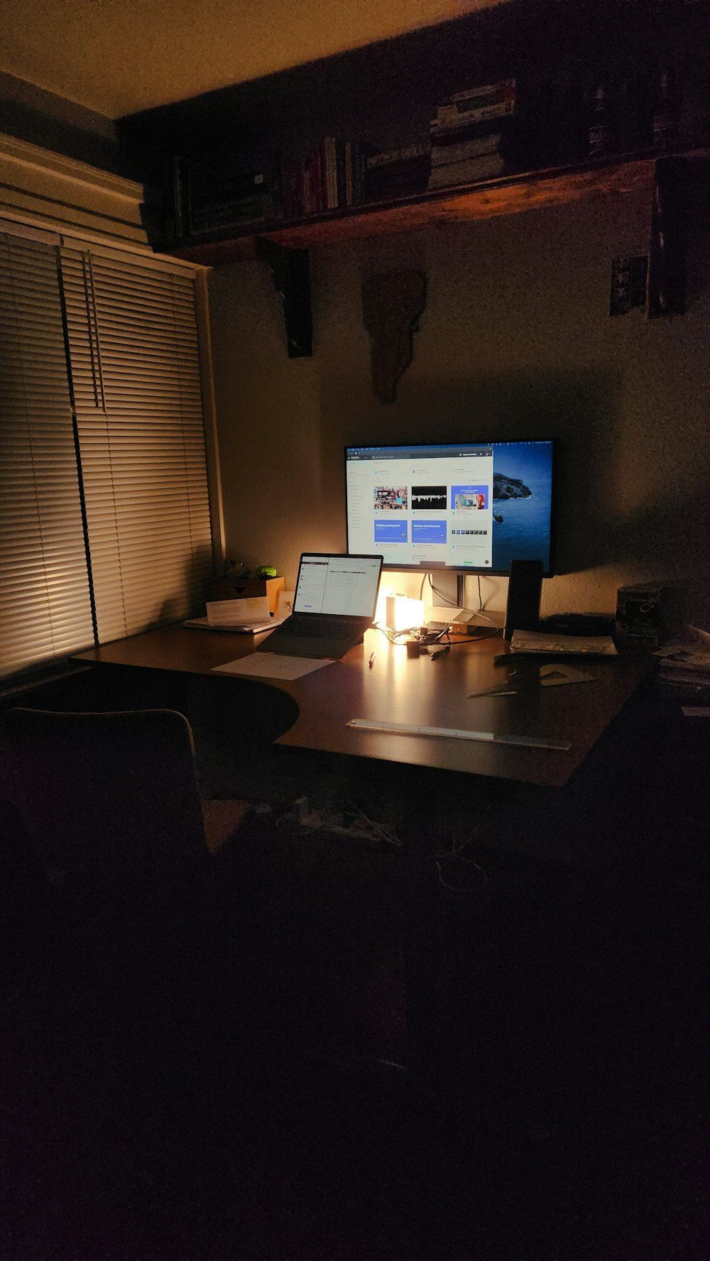 Dimly lit desk with a laptop, second monitor and desk lamp beside a window with blinds