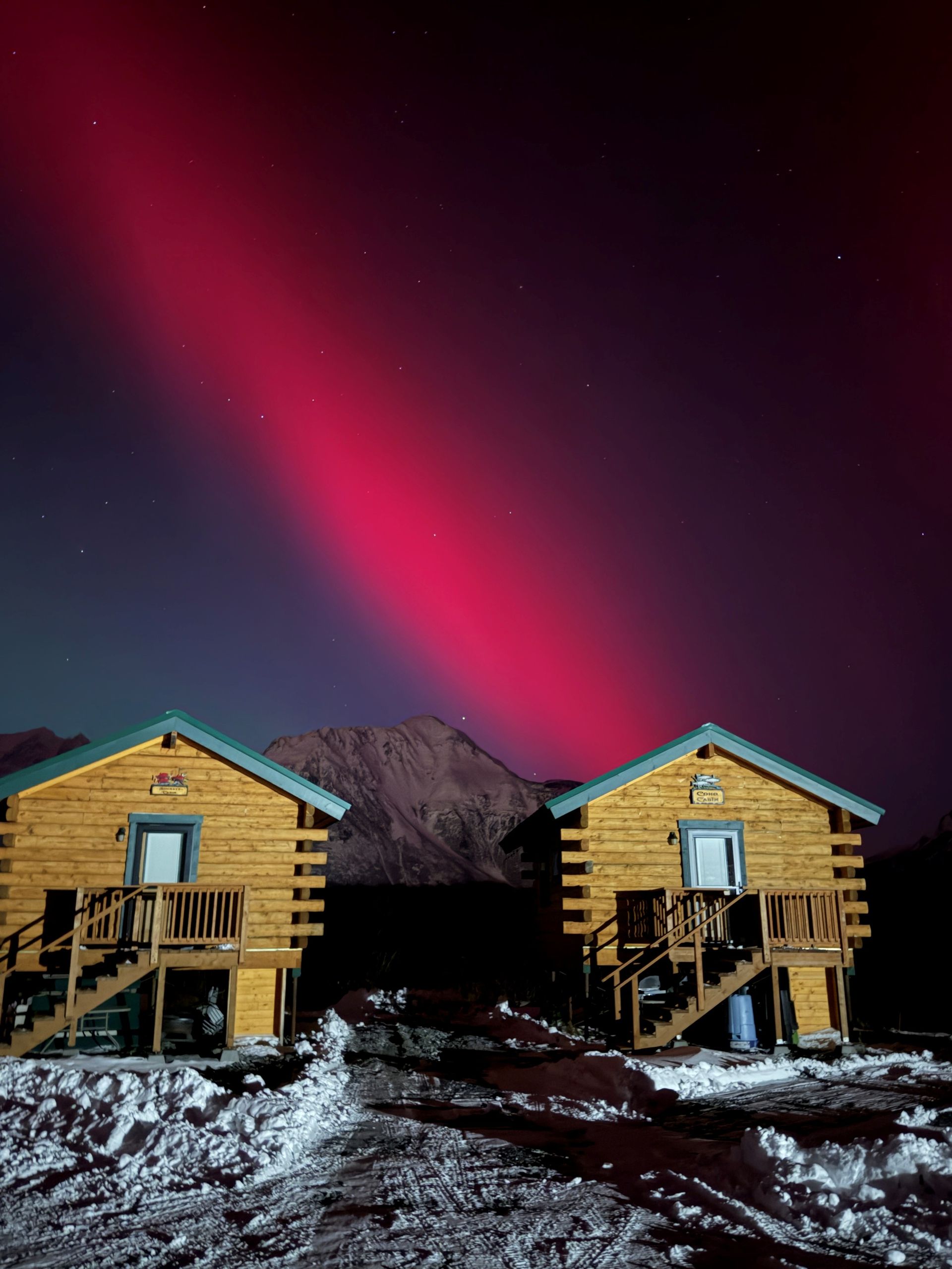 Two rustic wooden cabins sit in a snowy landscape under a dramatic, vibrant red aurora borealis in a dark night sky.