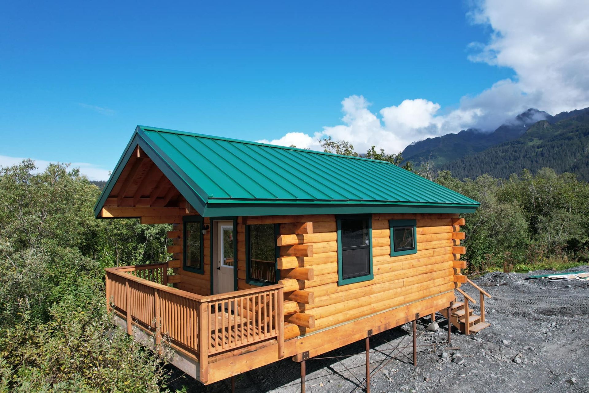 A small log cabin with a green roof is surrounded by trees and mountains.
