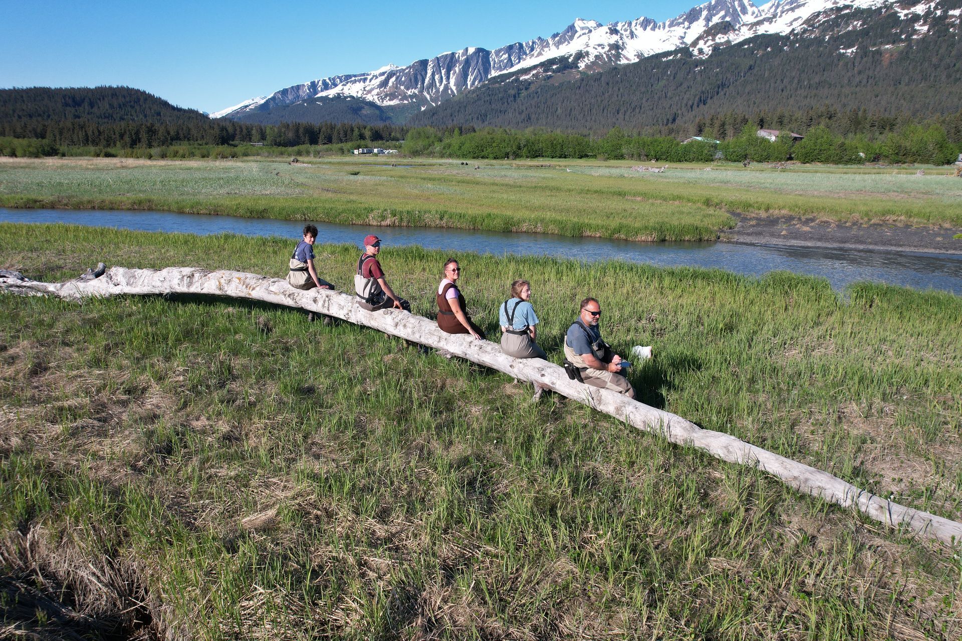 Five people sit in a row on a long fallen log in a grassy meadow, with a stream and snow-capped mountains in the background.