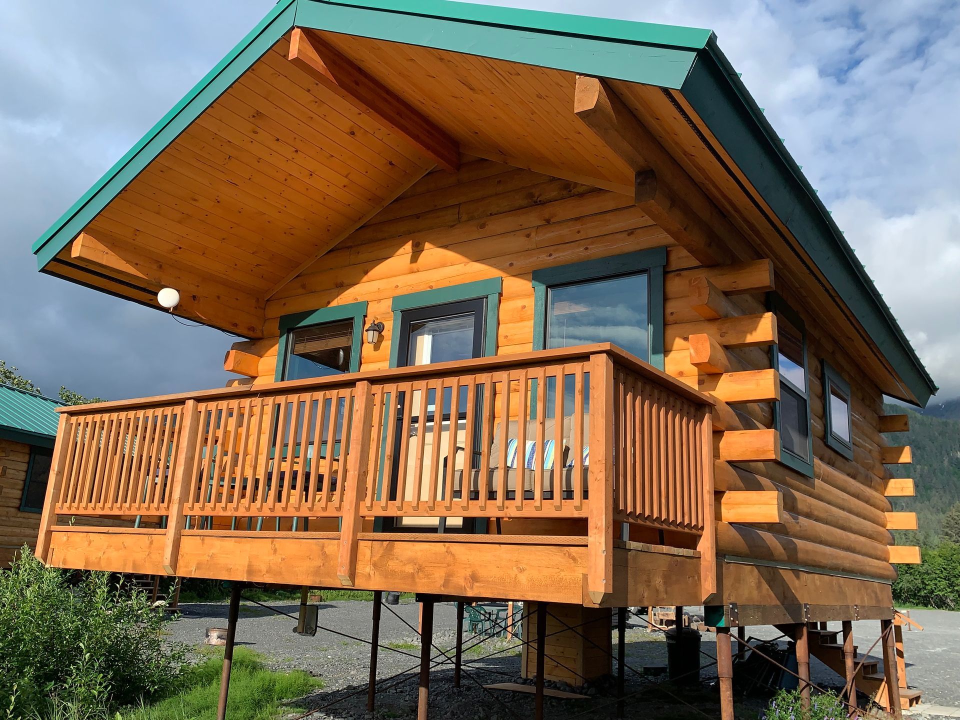 Log cabin with a wooden deck, raised on stilts, green roof.