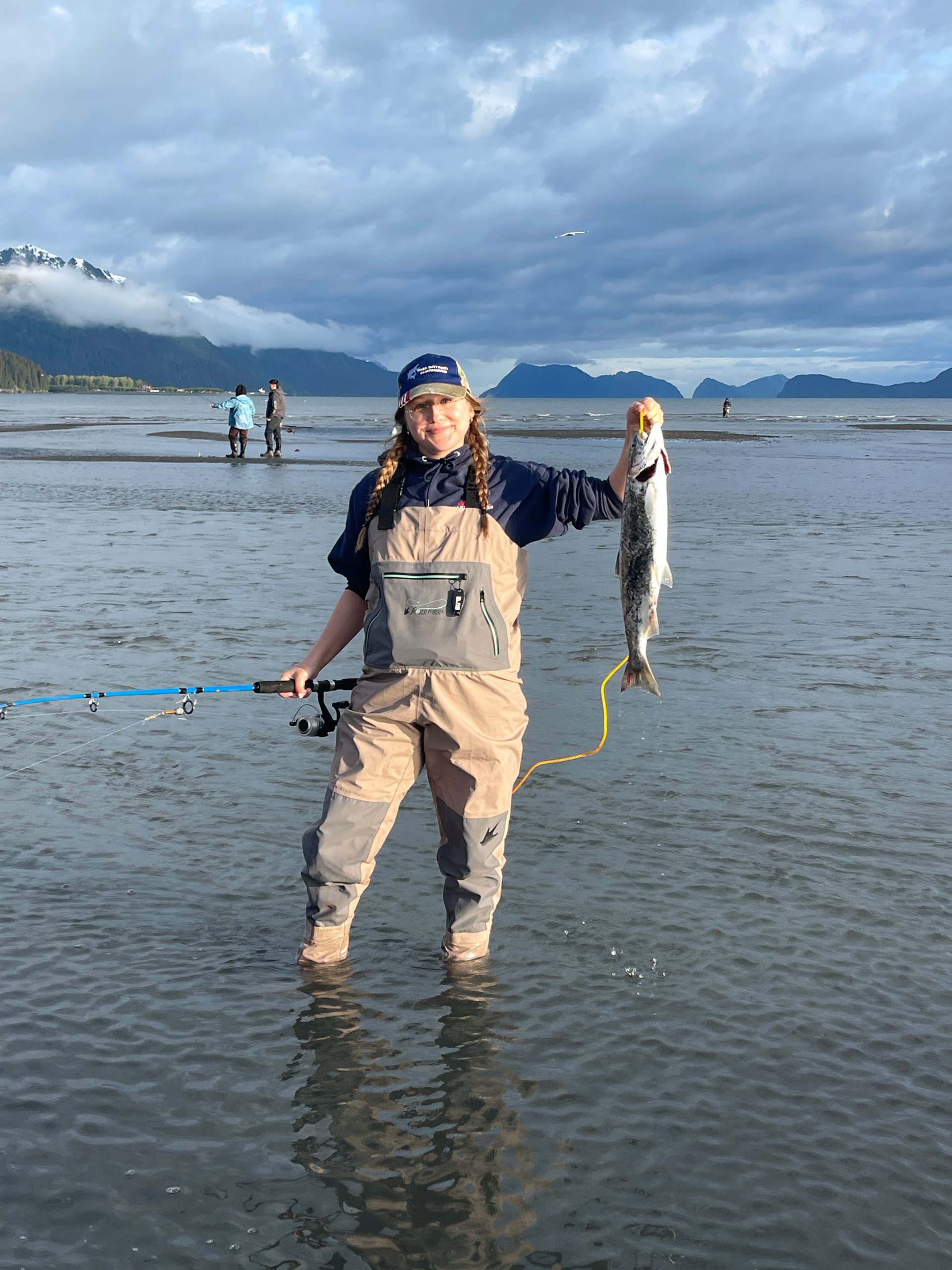 Woman in waders holding a fish, standing in shallow water, fishing rod in hand.