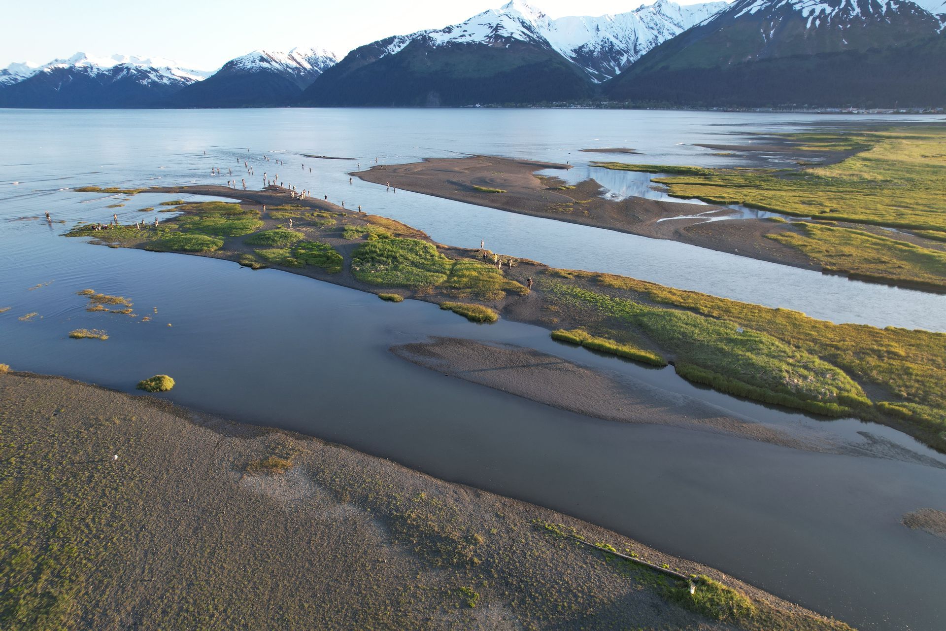 An aerial view of a river estuary meeting a calm bay, flanked by snow-capped mountains under a clear sky.