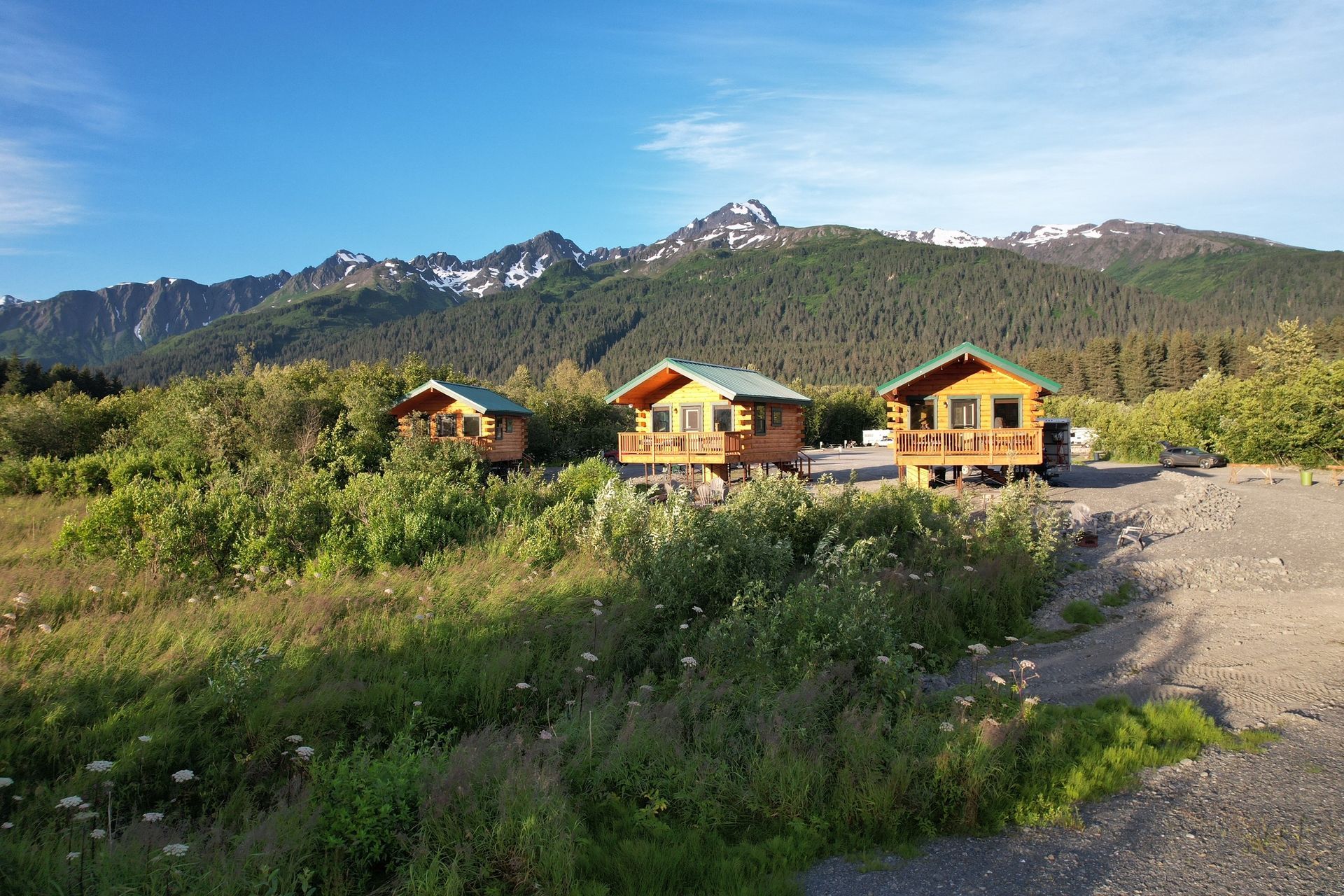 Three small wooden cabins sit in a grassy, gravelly field before a large, snow-capped mountain range under a blue sky.