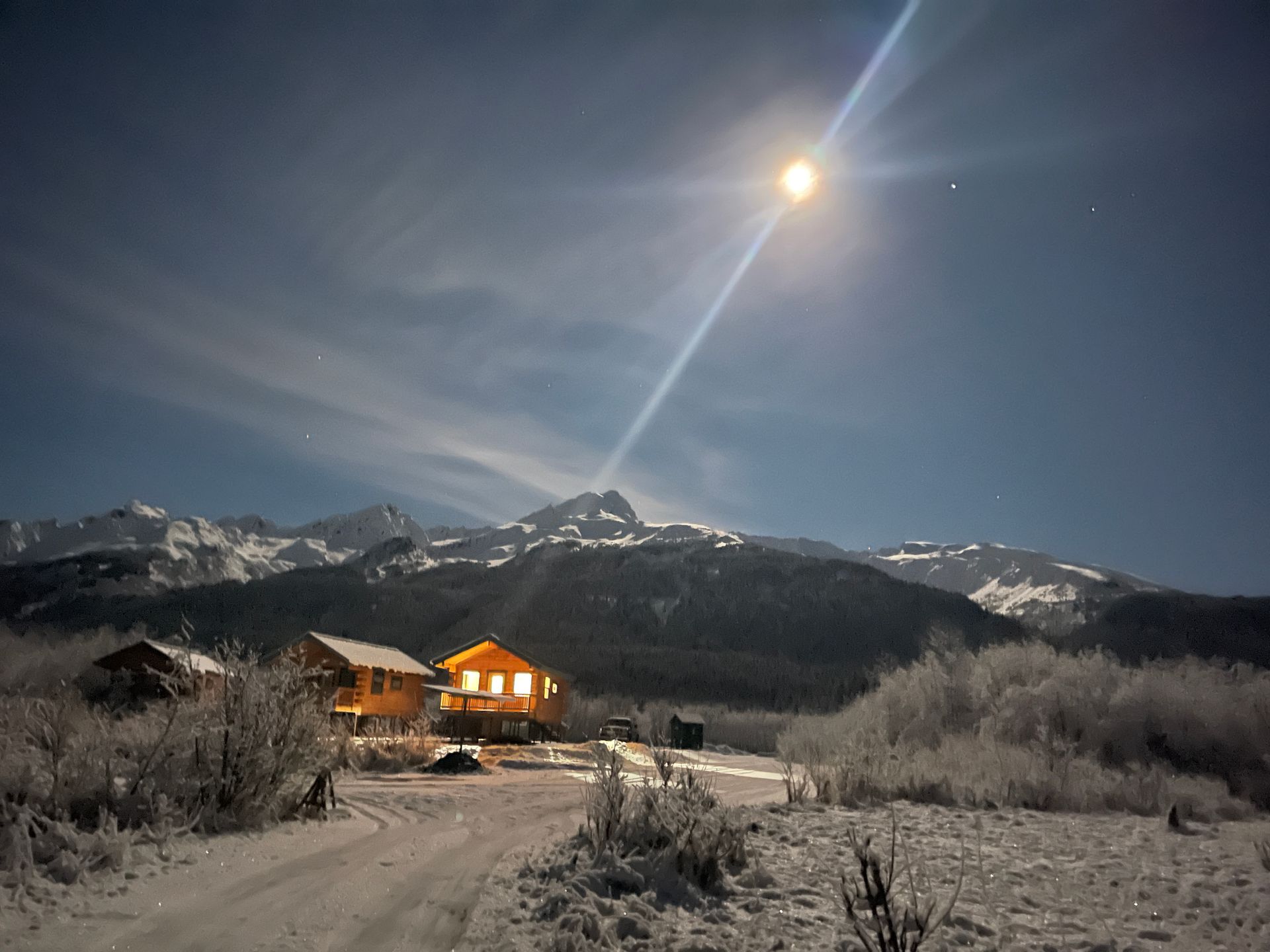 A bright moon shines over a snow-covered landscape with small, glowing wooden cabins nestled before jagged mountains.