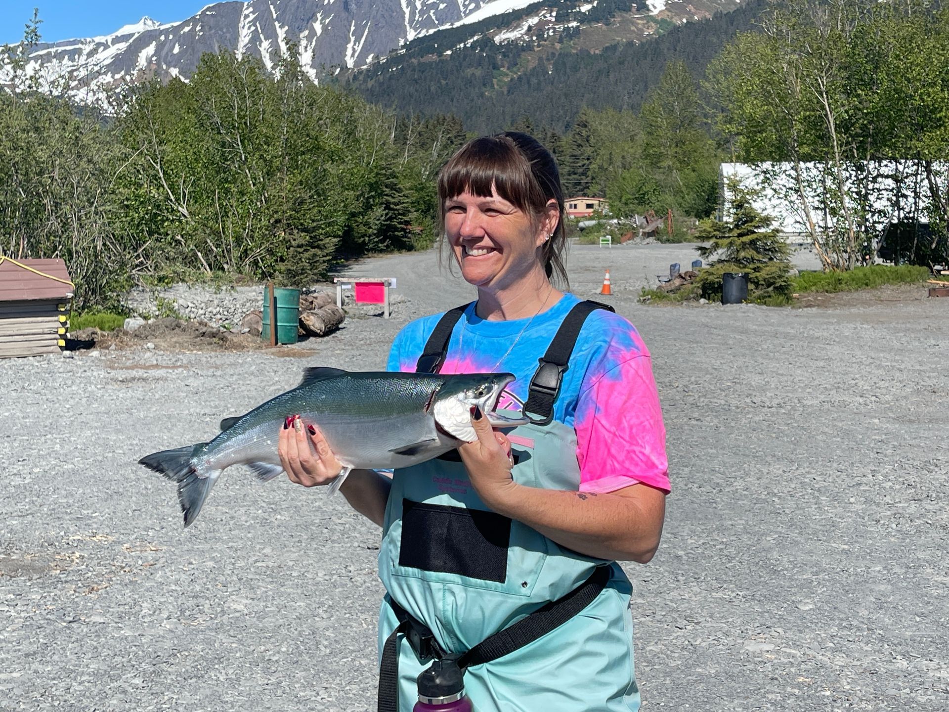 A smiling person in teal bib overalls and a colorful shirt holds a freshly caught silver fish in front of snowy mountains.