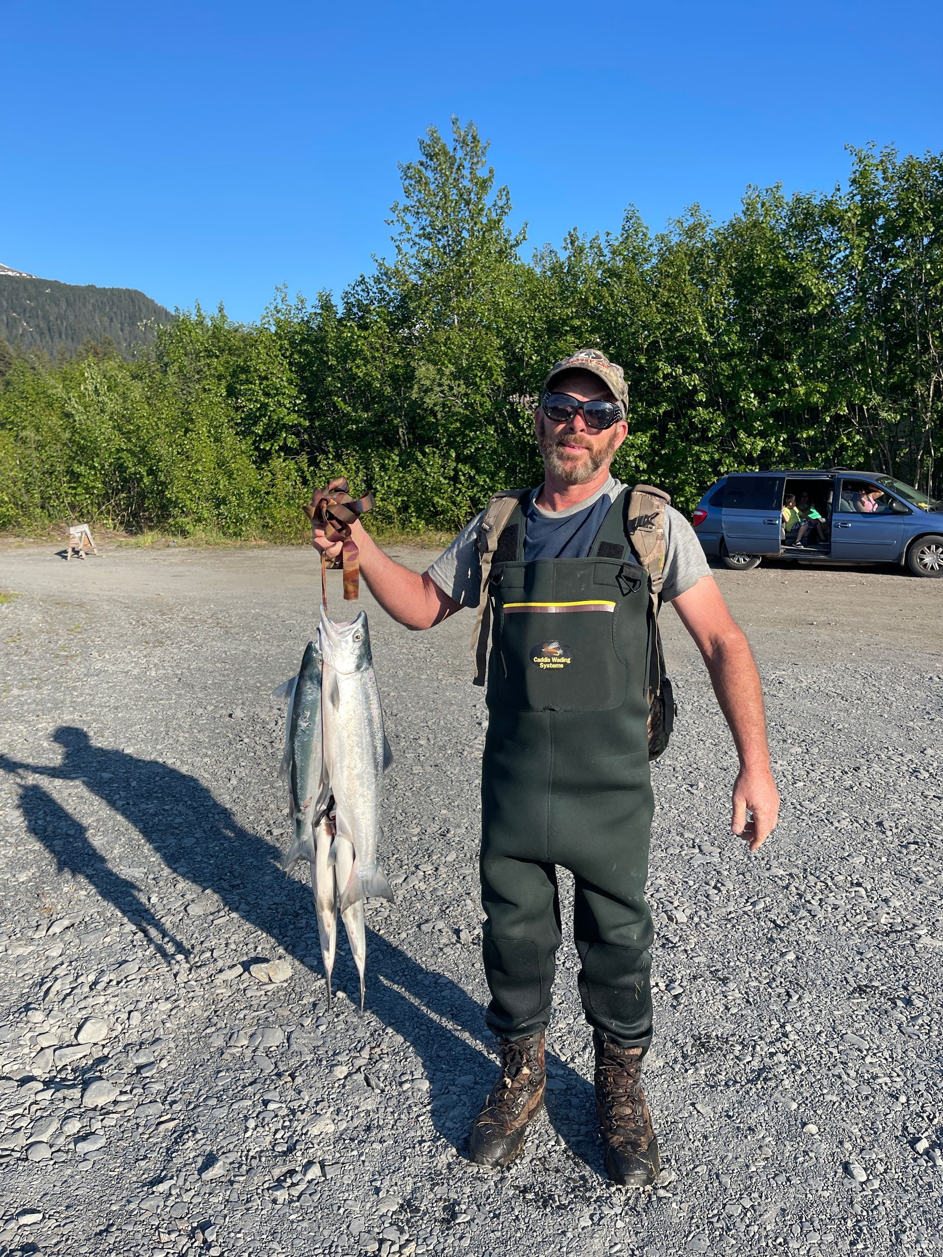 A person in waders stands on a rocky shore, holding a freshly caught fish against a backdrop of trees and a blue car.