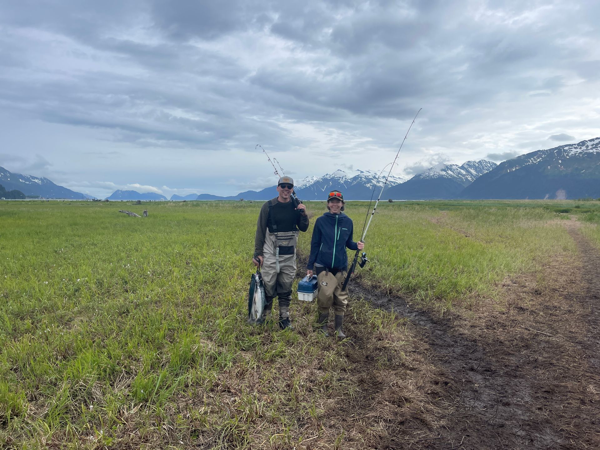 Two people in fishing gear with rods and a fish, walking through a green coastal field against a mountain backdrop.