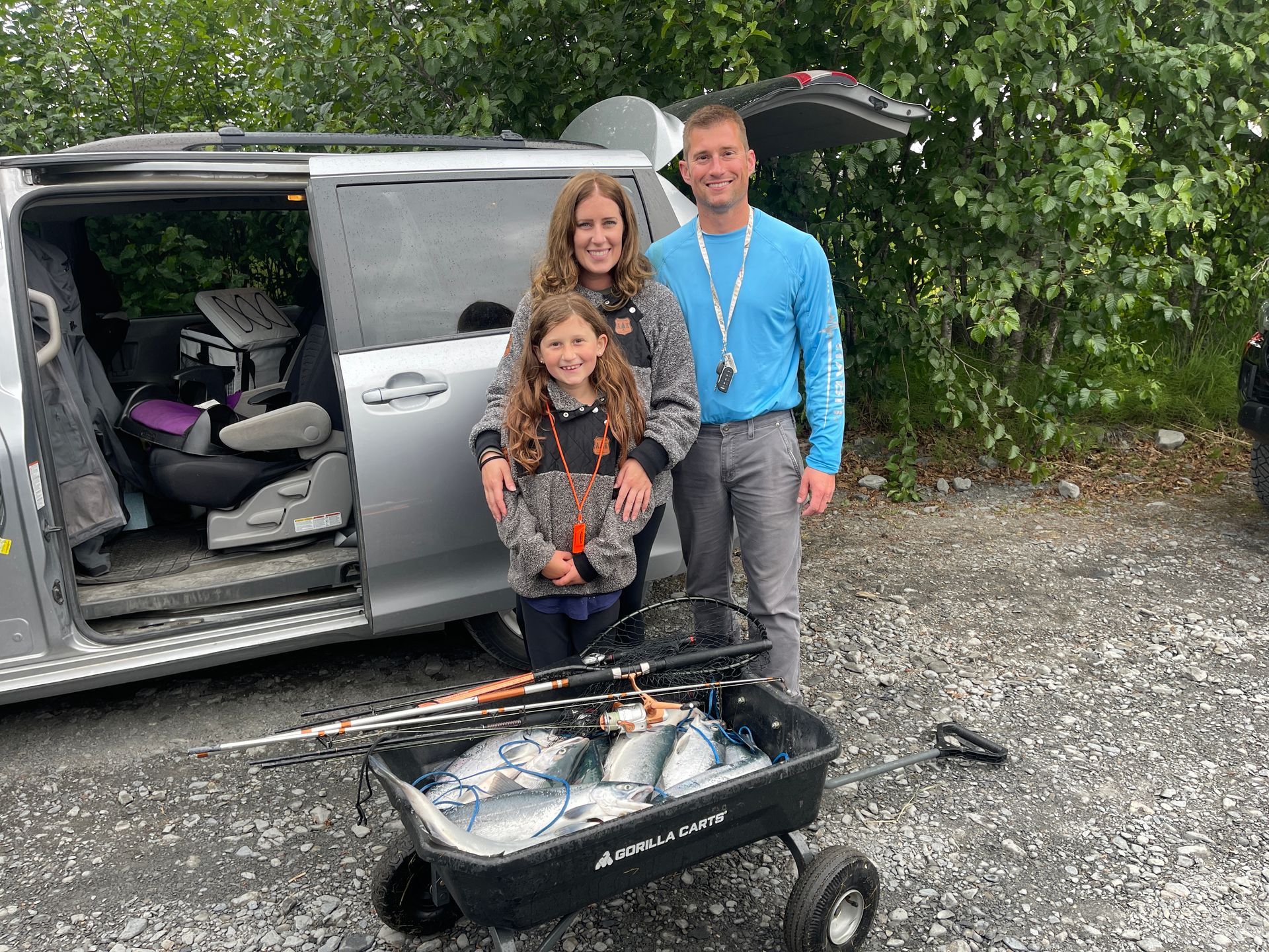 A family stands next to a minivan with a wagon filled with fresh fish in a gravel lot surrounded by trees.